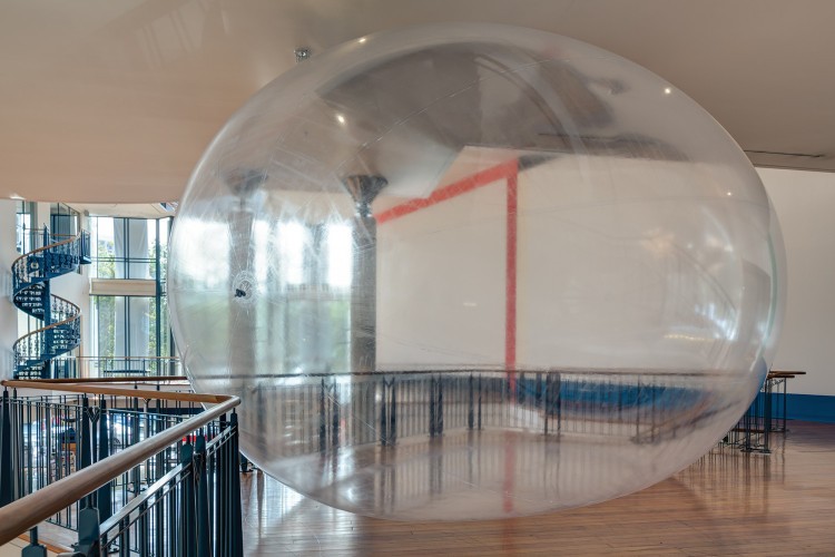 Large translucent inflated orb sandwiched between the floor and ceiling, and next to handrails.