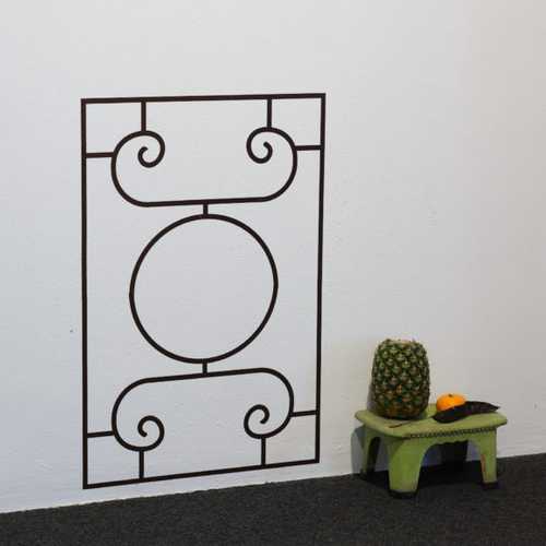 A decorative grille is painted onto a wall, next to a green low stool with a pineapple and mandarin sitting on top.