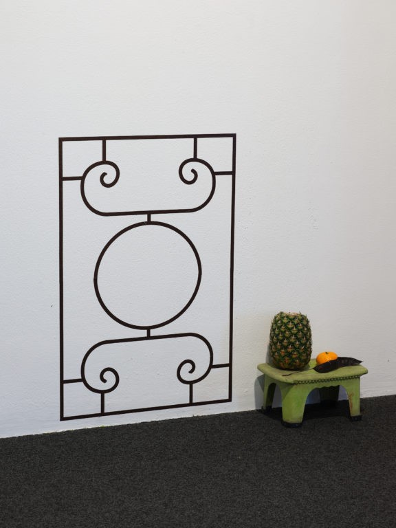 A decorative grille is painted onto a wall, next to a green low stool with a pineapple and mandarin sitting on top.