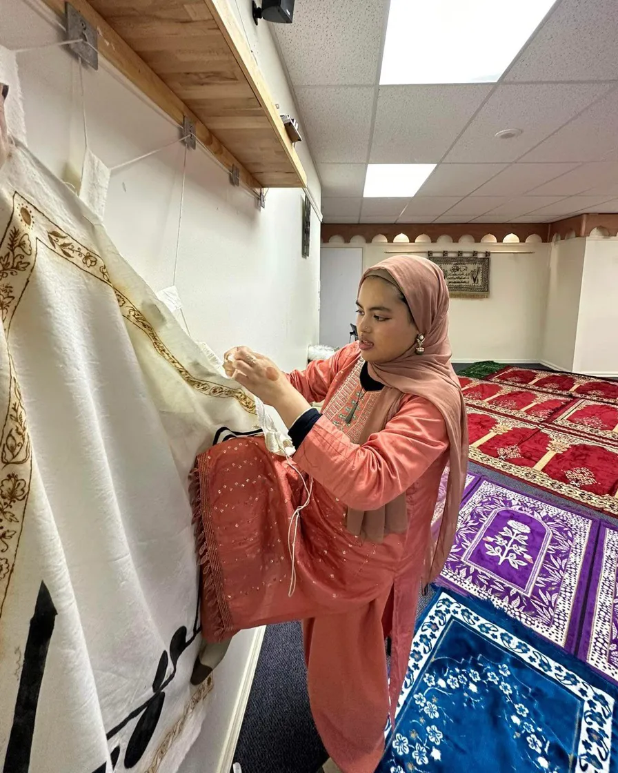 A person in hanging a large piece of embroidered fabric to a wall.