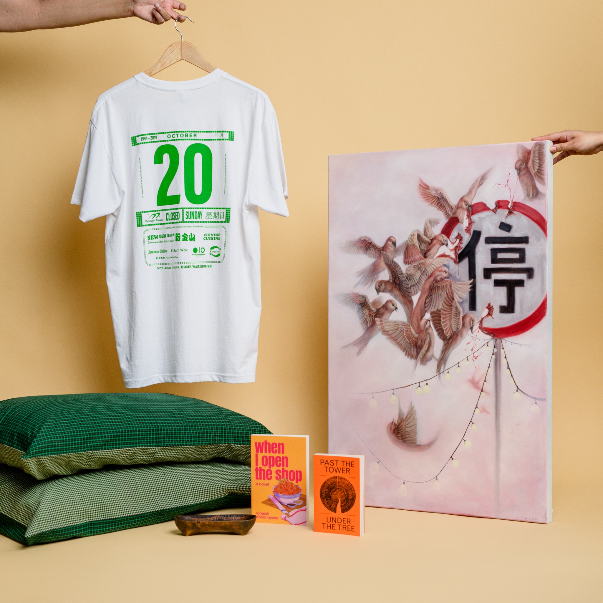 A white t-shirt with green calendar design, two green pillows, two books, a wooden tray and a large painting with birds attacking a sign