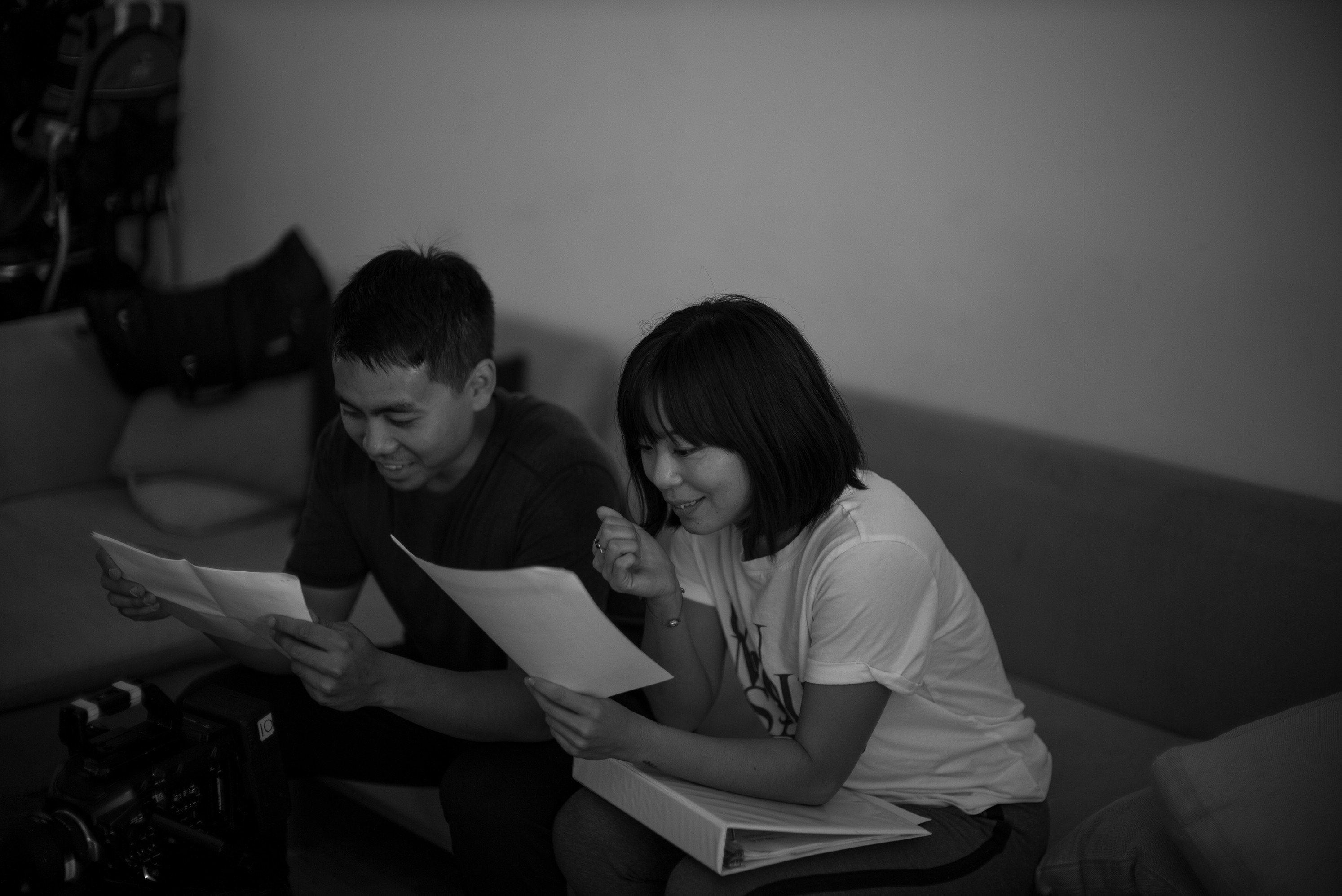 Black and white behind-the-scenes image of a film shoot, showing a man and a woman sitting on a sofa, with each holding a film script.