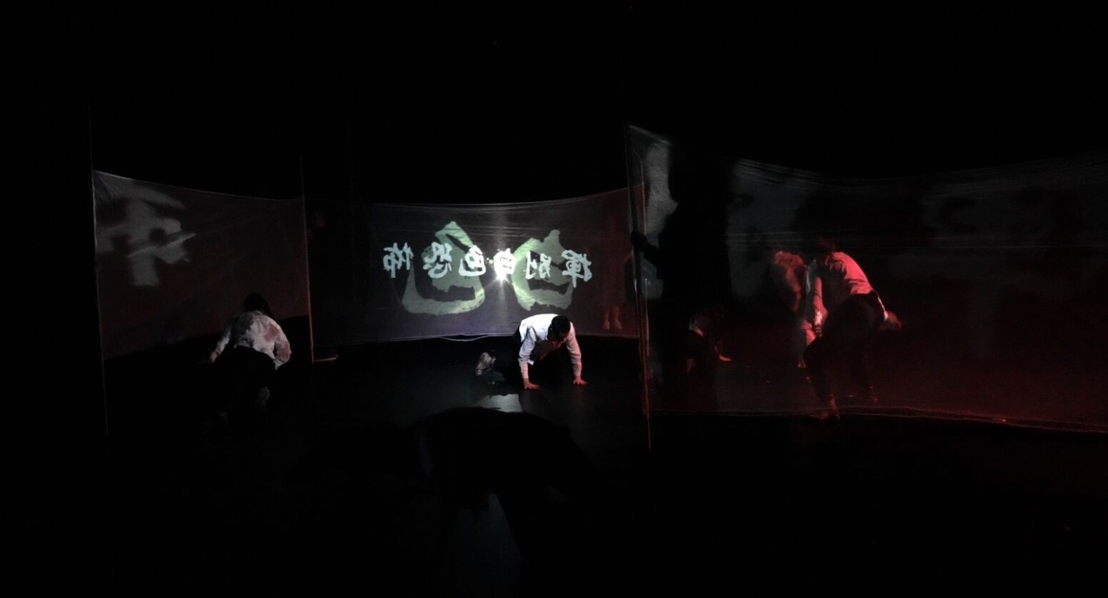 Performers dancing in a large dark space. Projections of words from the 1991 Taiwanese protest shining through white meshes and landing on the dancers' body