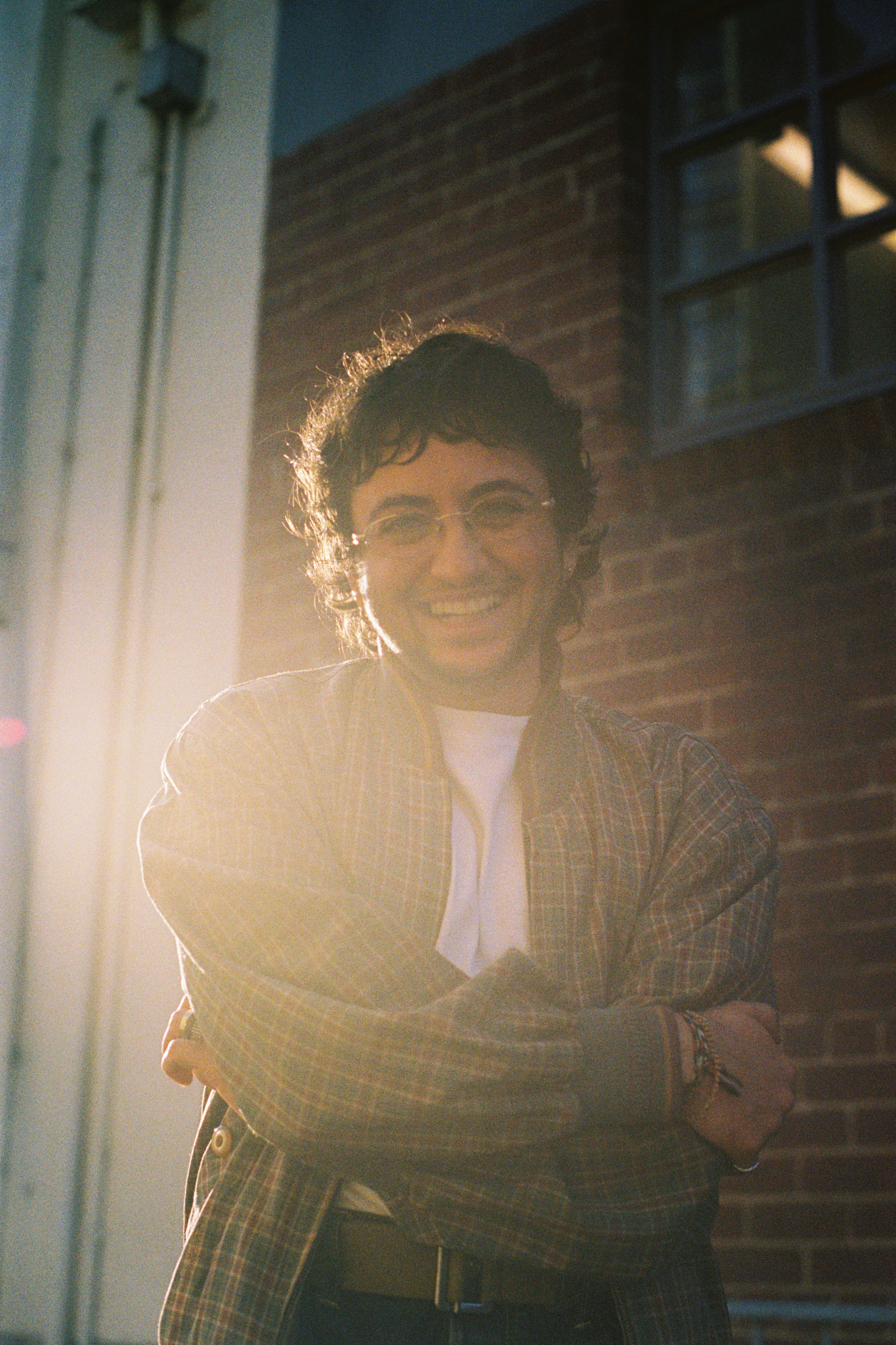 A man in a check jacket and curly brown hair with glasses smiles at the camera. Sunlight bursts around him.