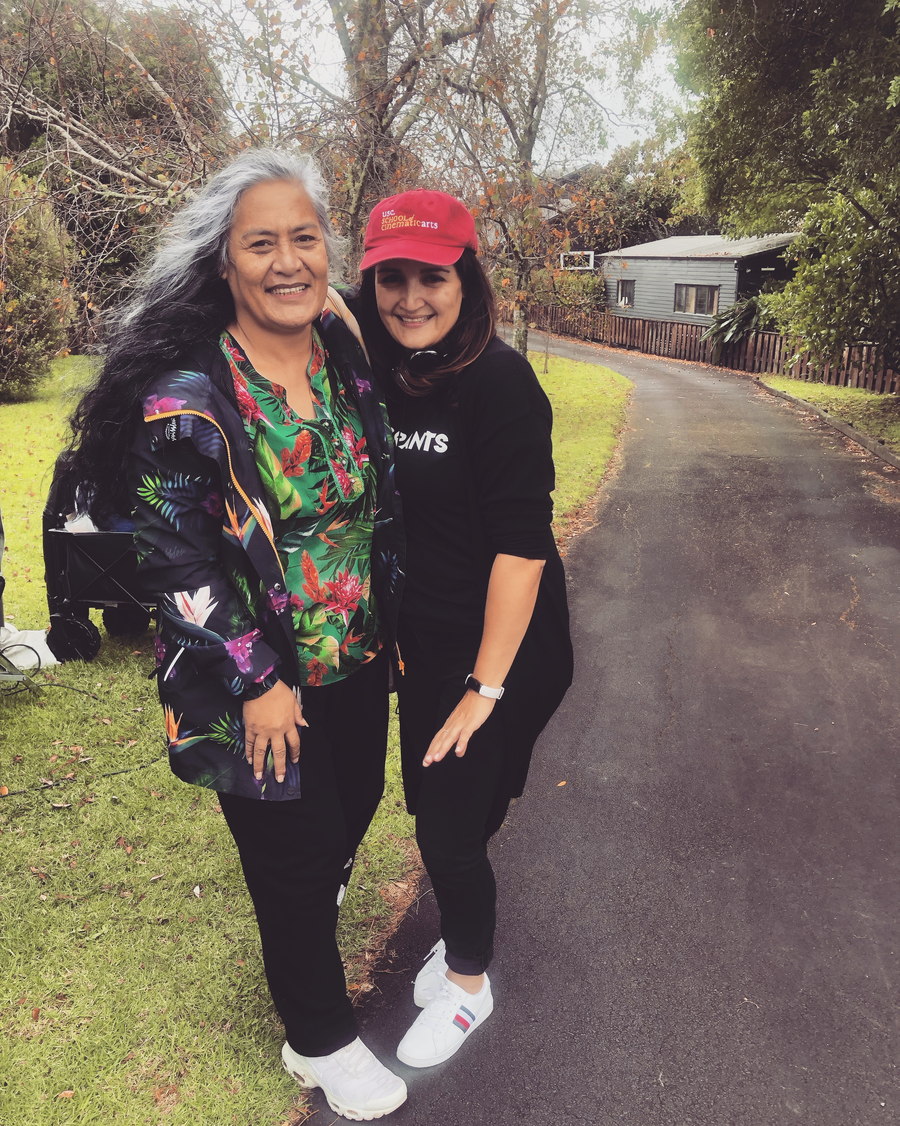 Two women smile and pose together on a tree-lined path, one wearing a bright floral shirt and the other in a red cap and black outfit.