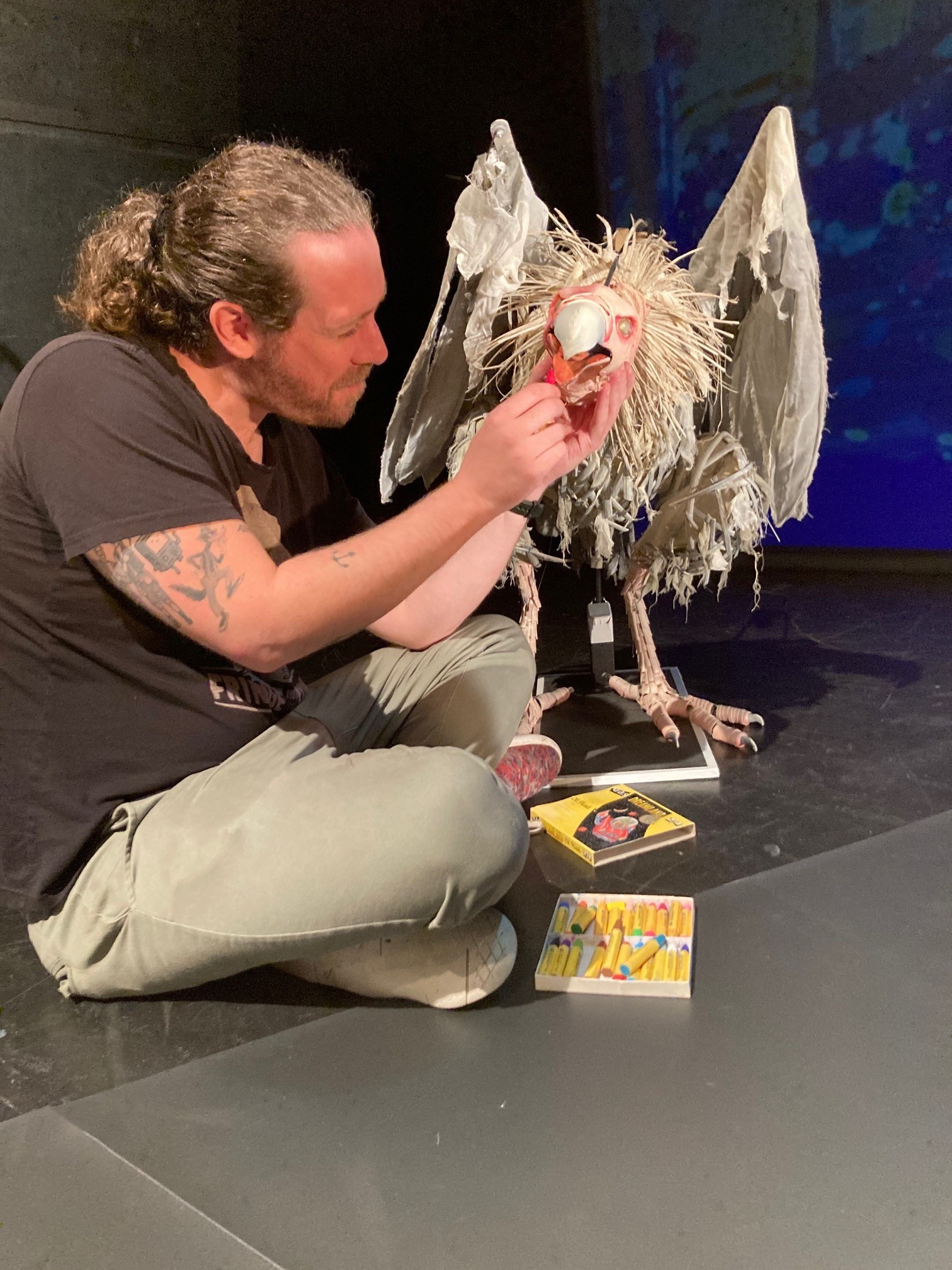 A man sitting cross-legged on the ground holding open the mouth of a vulture puppet