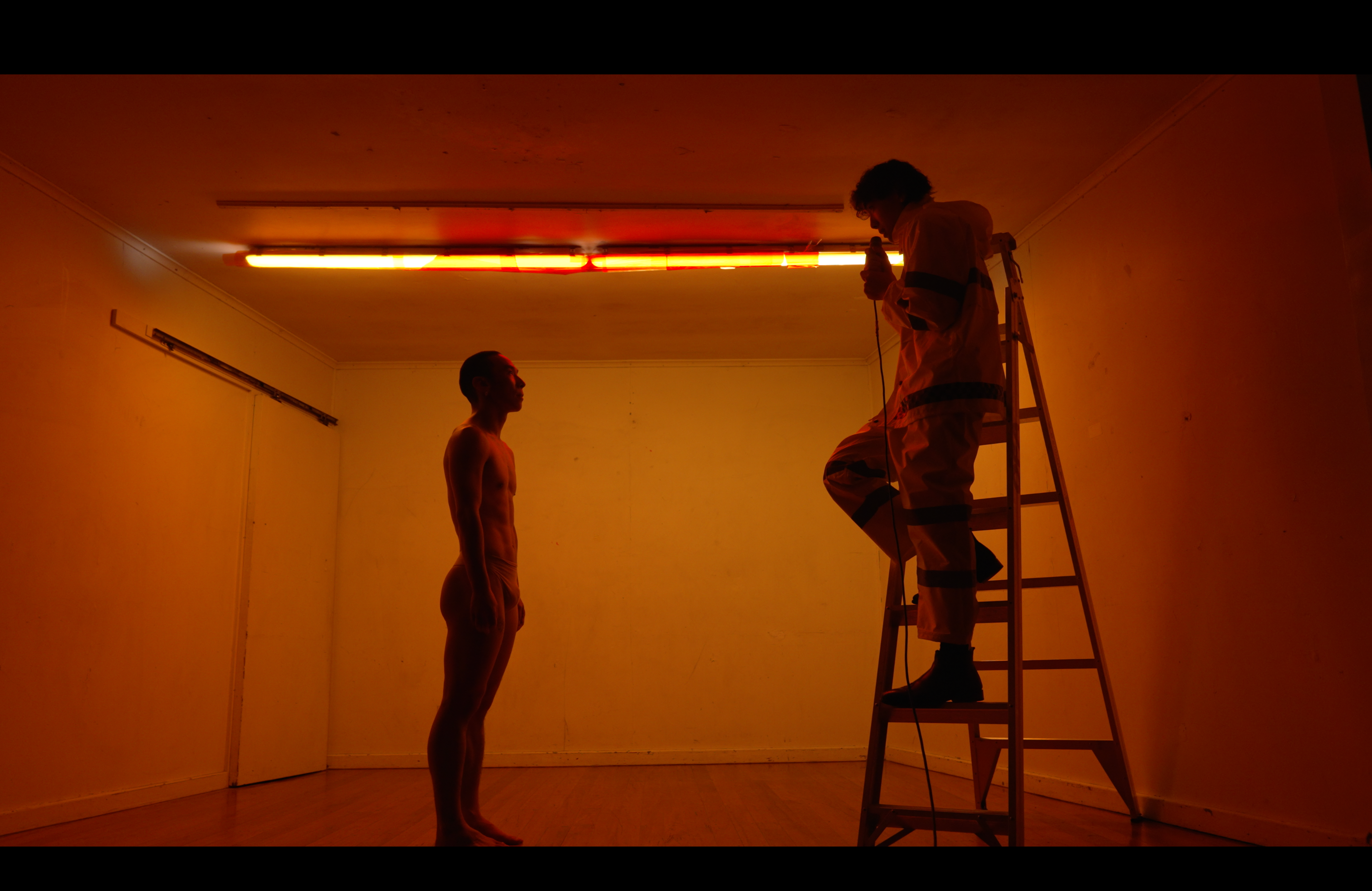 In a small orange-lit room, a shirtless man gazes up at a man in a hazmat suit standing on a ladder