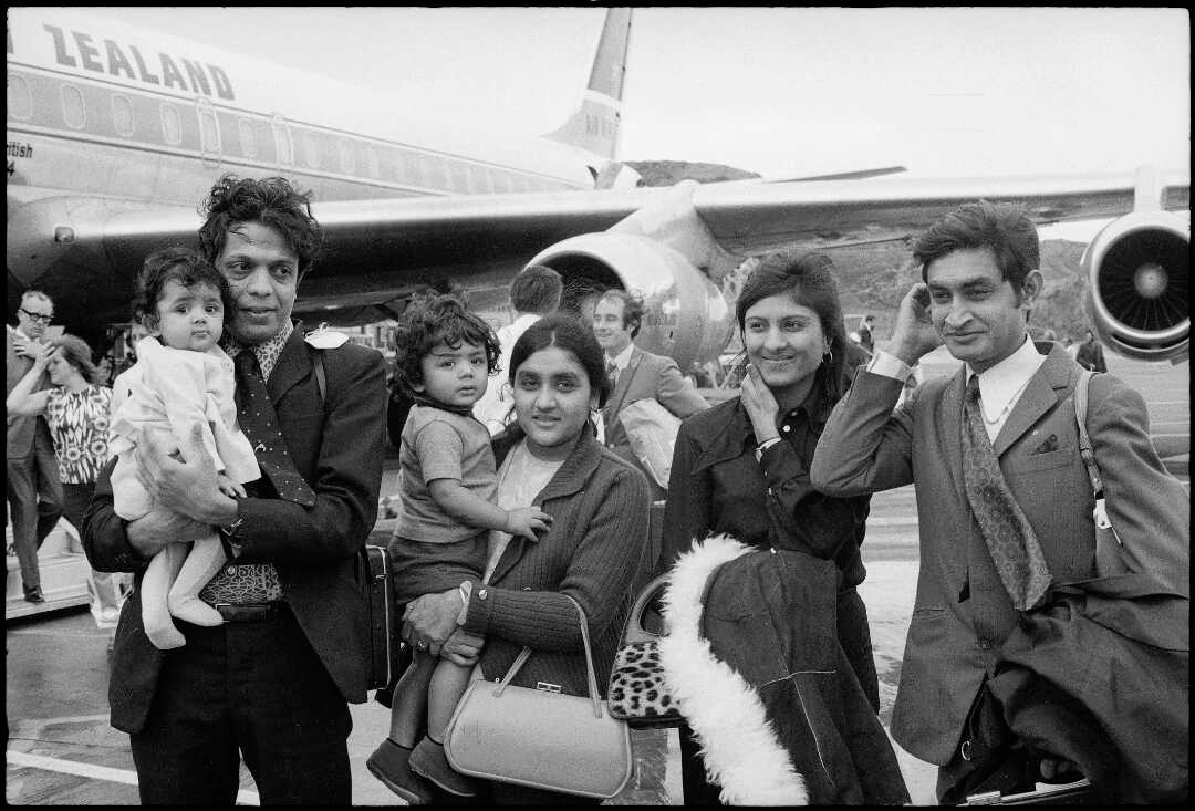 Four Indian adults, two holding young children, are smartly dressed standing on the tarmac in front of an Airt New Zealand plane.