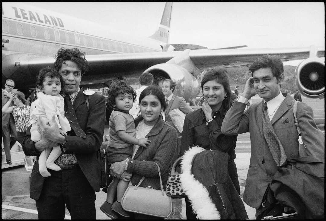 Four Indian adults, two holding young children, are smartly dressed standing on the tarmac in front of an Airt New Zealand plane.
