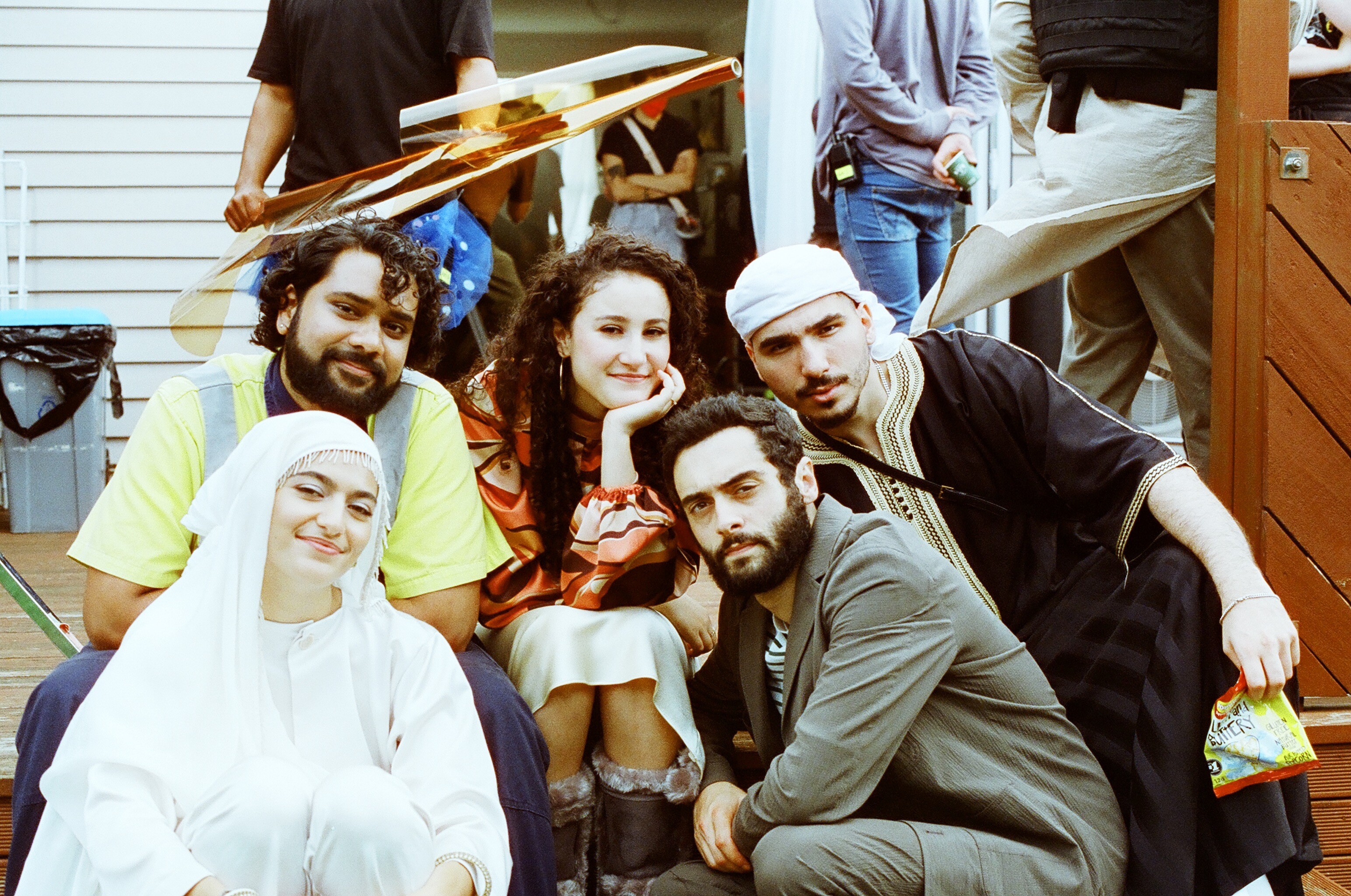 A group of five cast members sit closely together on a film set, smiling and dressed in a mix of costume and casual wear.