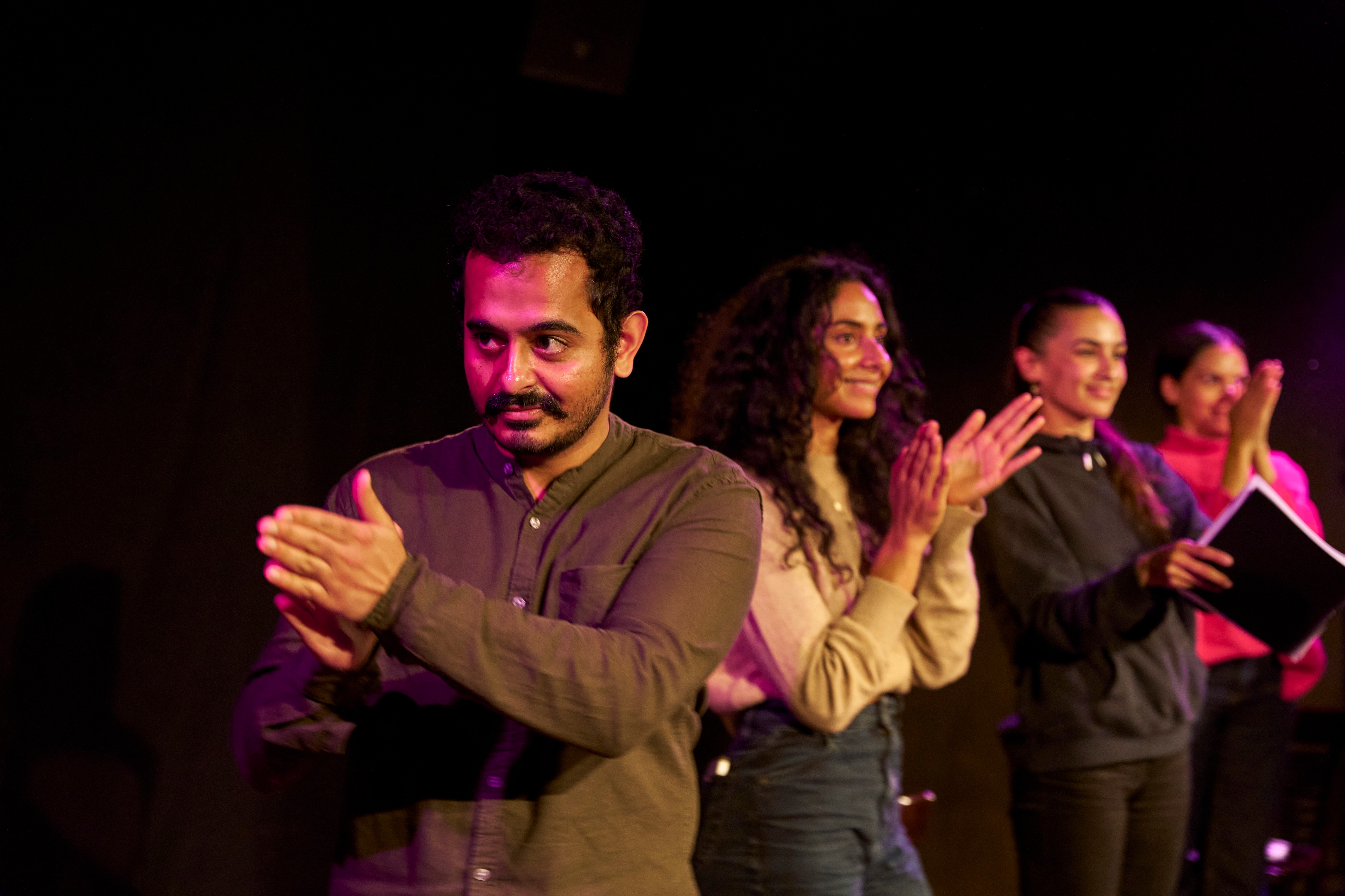 A line of actors all clapping. One man is looking to his right at someone while the others are looking into the audience.