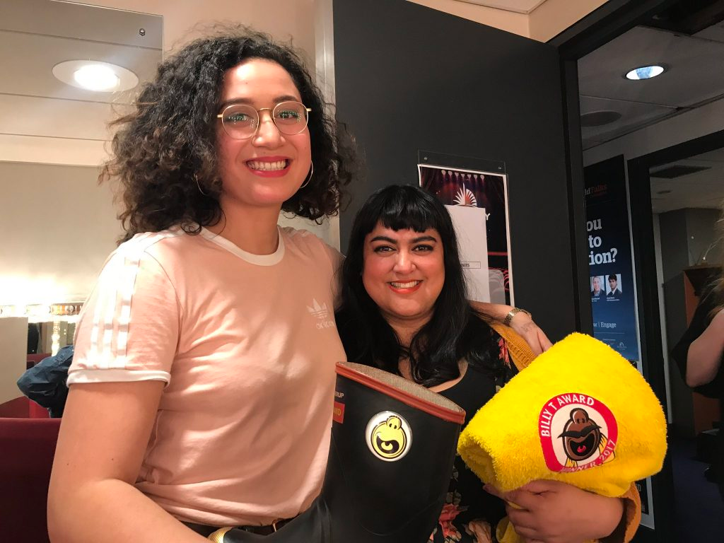 Two women smile at the camera together. One is tall with curly black hair and glasses, wearing a dusky pink adidas top and holding a 'gumboot' award, and the other is in black holding the Billy T towel