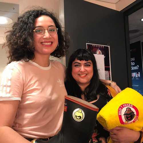 Two women smile at the camera together. One is tall with curly black hair and glasses, wearing a dusky pink adidas top and holding a 'gumboot' award, and the other is in black holding the Billy T towel