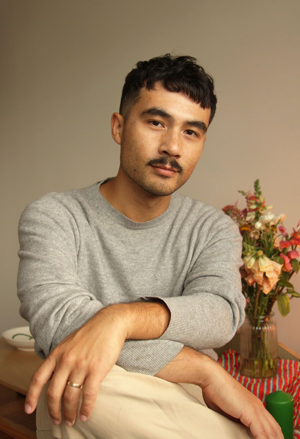 Close-up of a man with black hair and a moustache wearing a grey sweater with a vase of flowers in the background