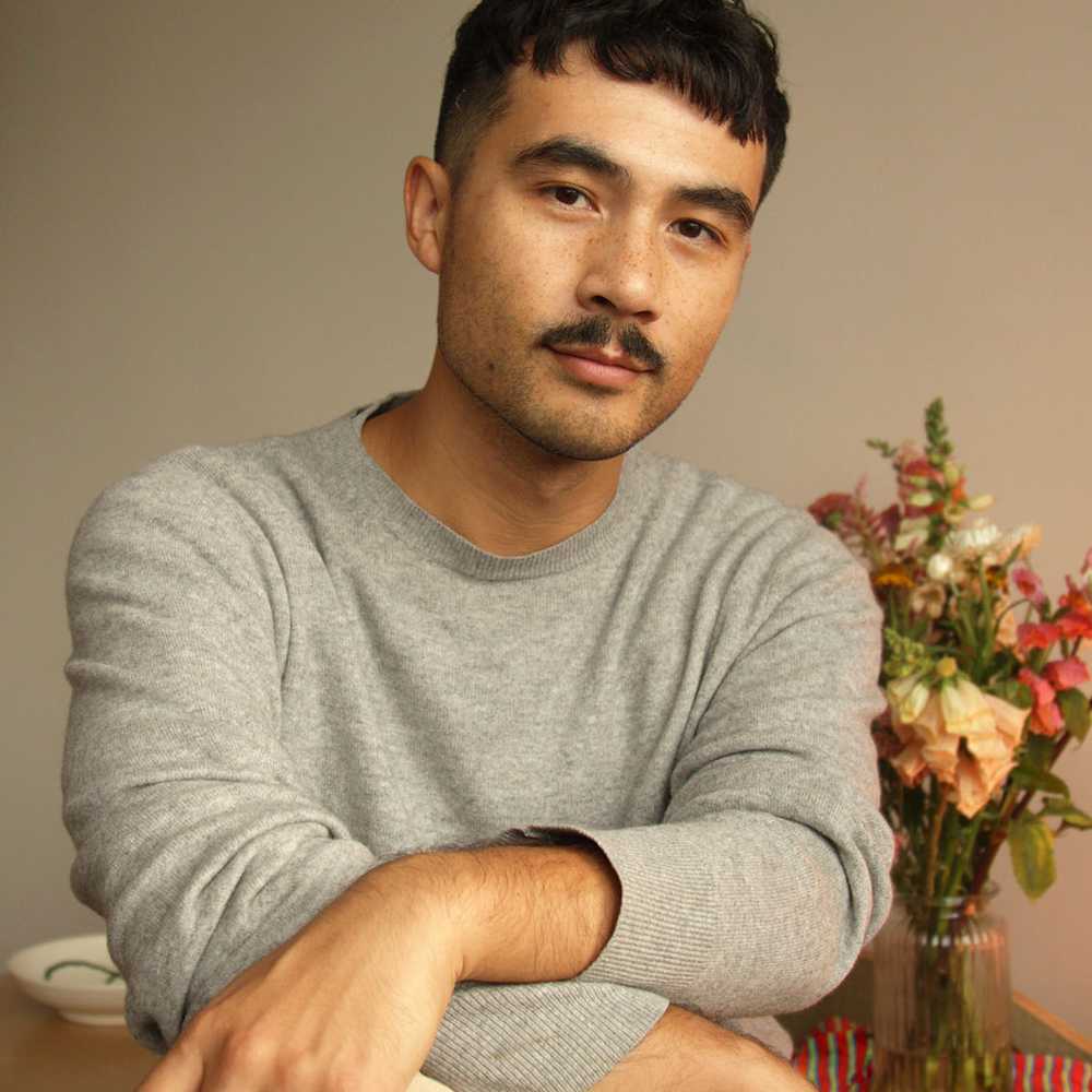 Close-up of a man with black hair and a moustache wearing a grey sweater with a vase of flowers in the background