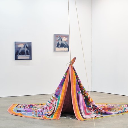 A quilt being raised off the ground with a string. Paintings of horses are in the background.