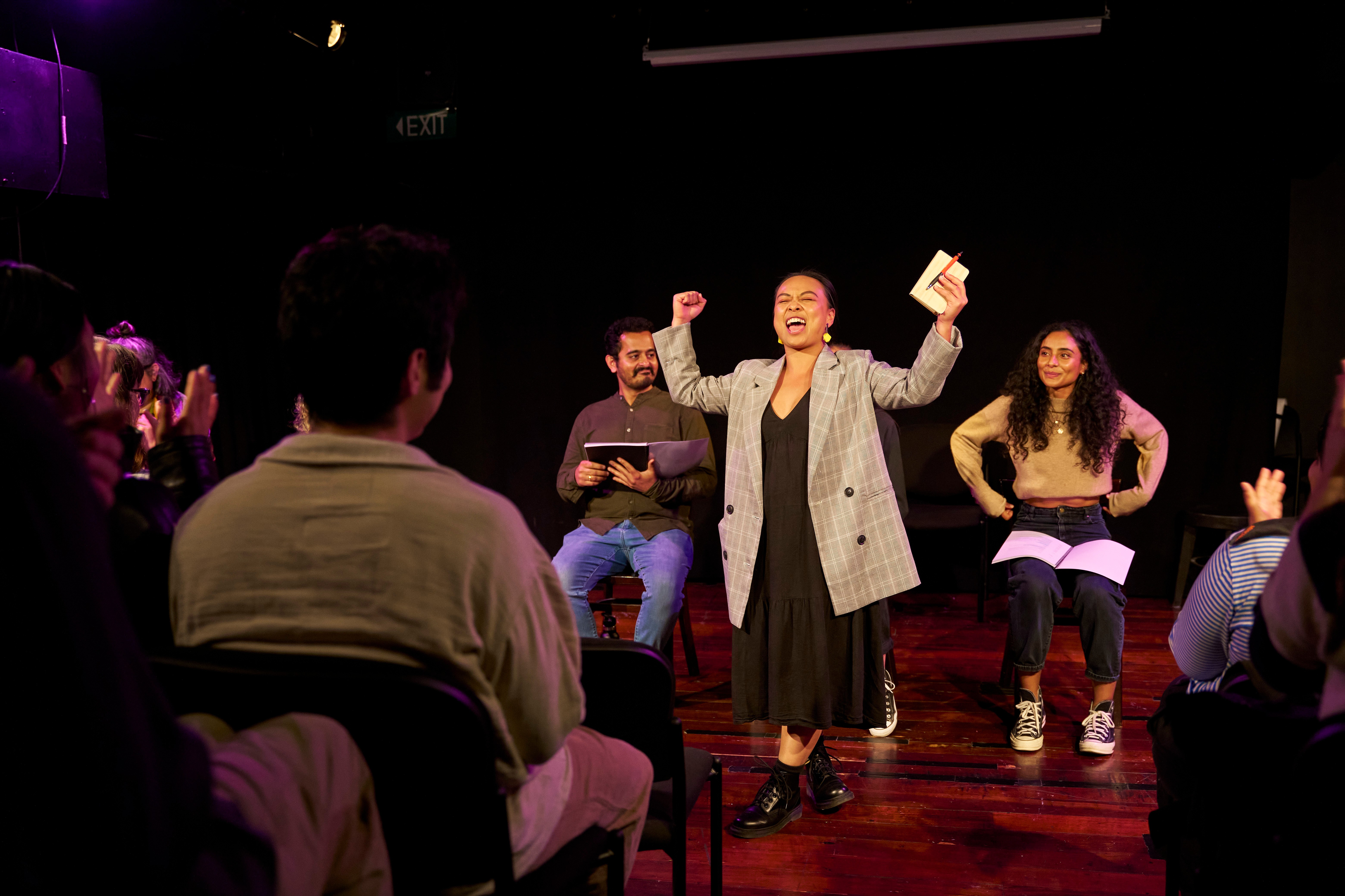 A woman in a grey blazer waves her arms in the air in a triumphant move, while people sit behind her with scripts in their lap