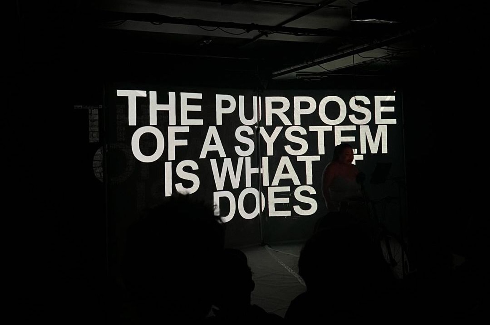 Projection on stage that reads 'the purpose of a system is what it does'.