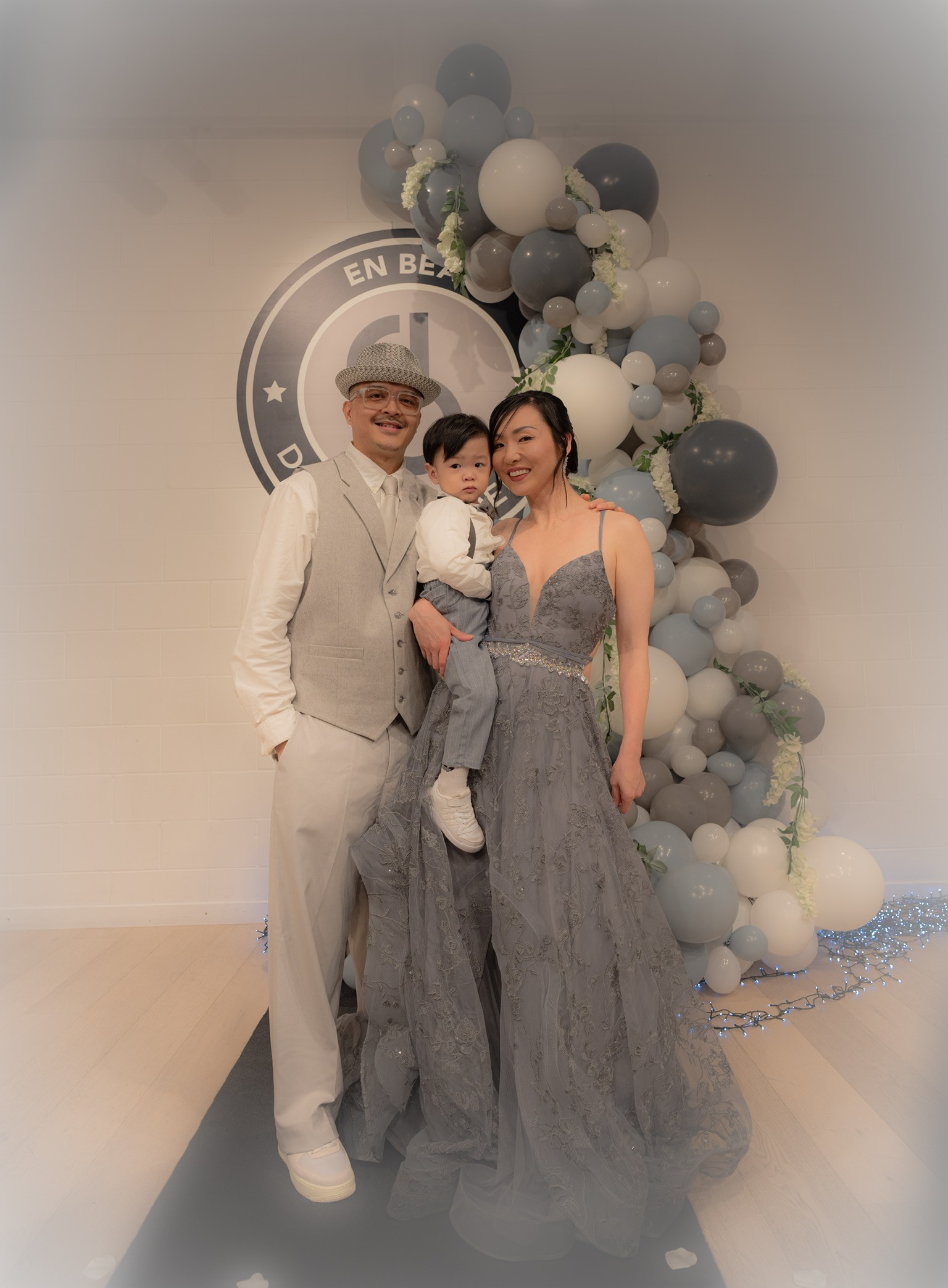 A man in a grey suit stands next to a woman in a grey gown. She is holding a baby in formal wear. A grey balloon structure is behind them with the logo of the academy