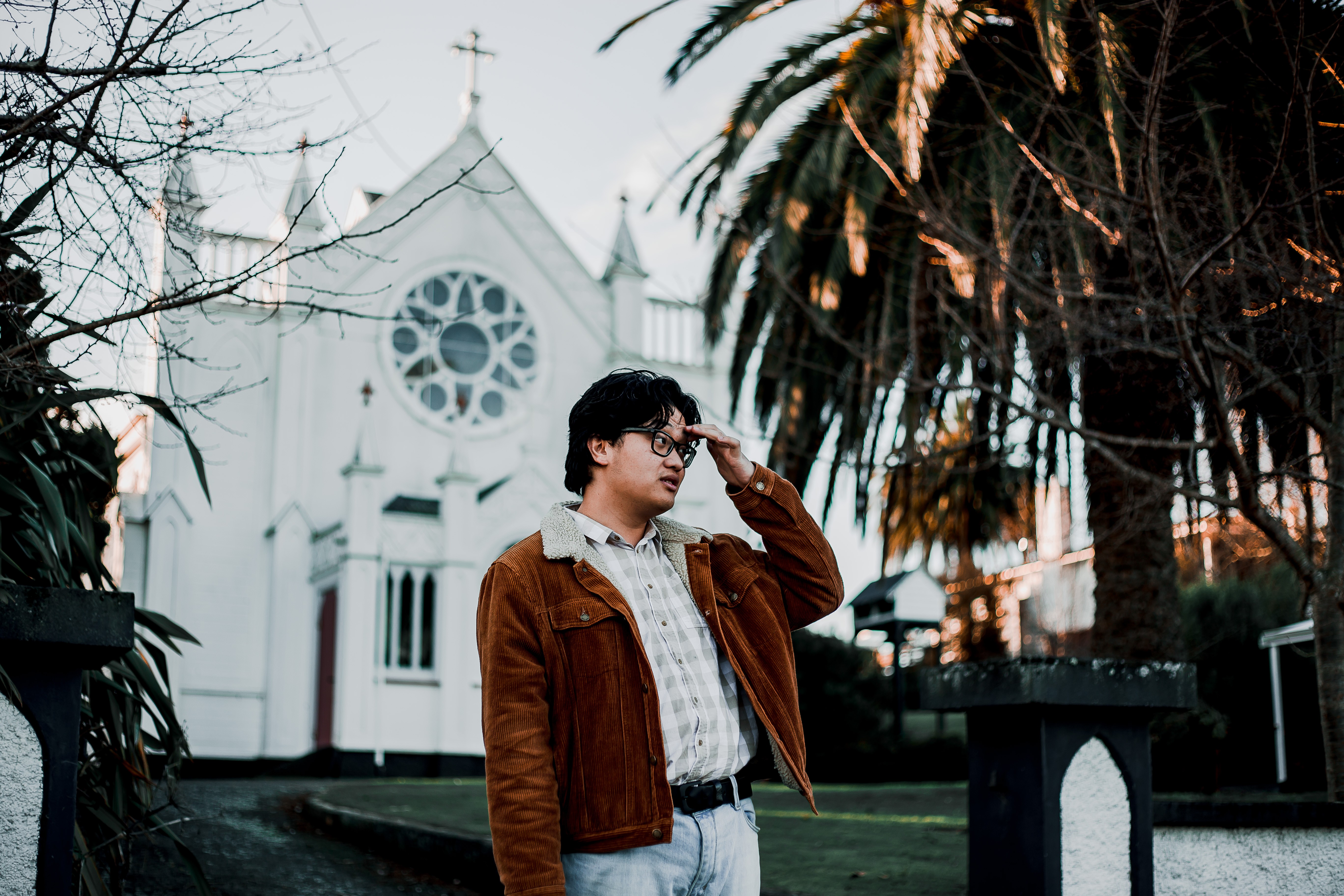 A man in a brown coat with dark wavy hair and glasses stands in front of a white church.