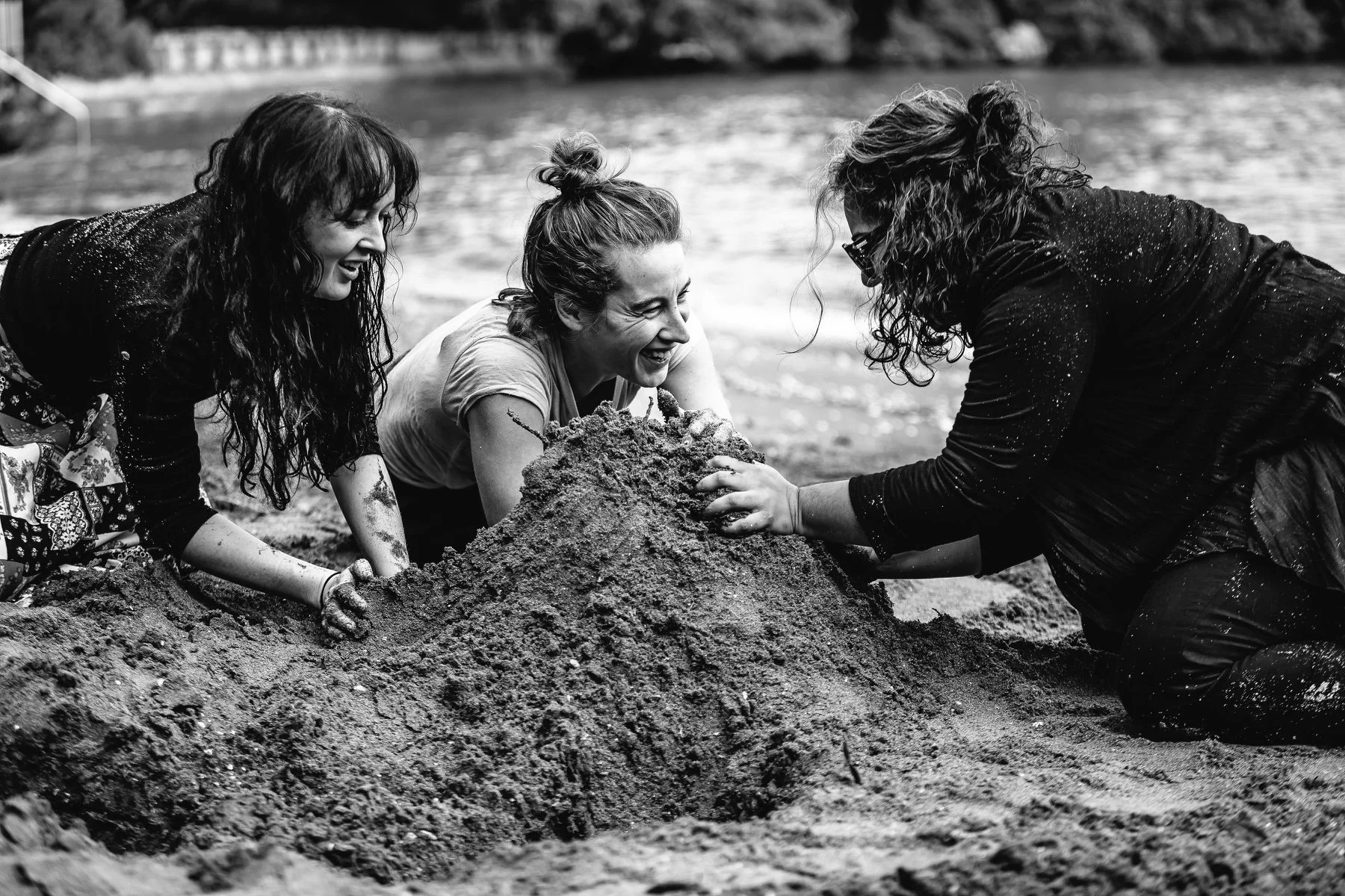 A black-and-white photo of three women heaping piles of sand on the beach and laughing