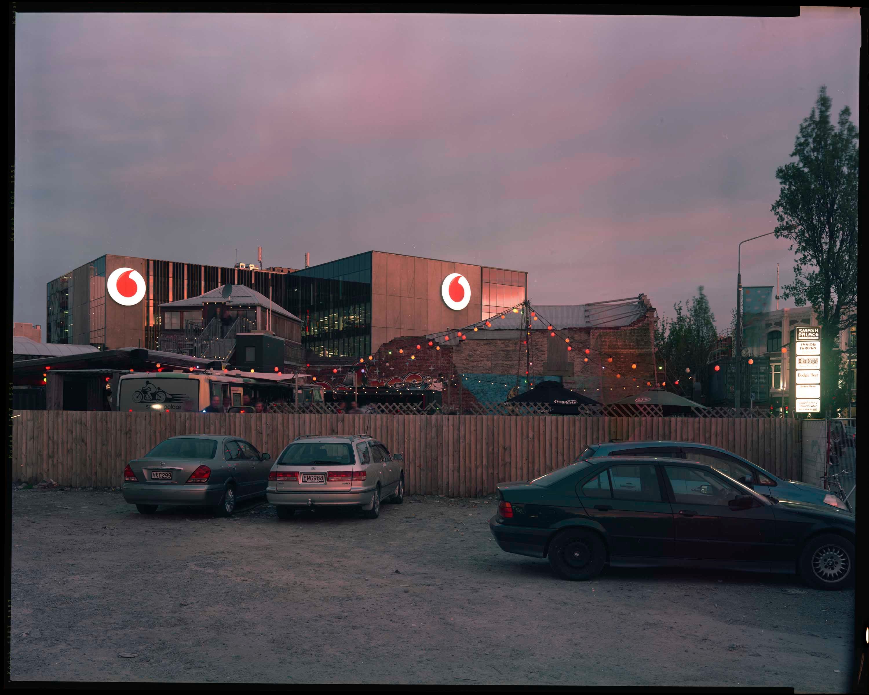 Four cars in a gravel carpark behind which a new building is visible. The lighting is pink and dusky, with a string of colourful lights and umbrellas between the carpark and new building indicating a makeshift bar.