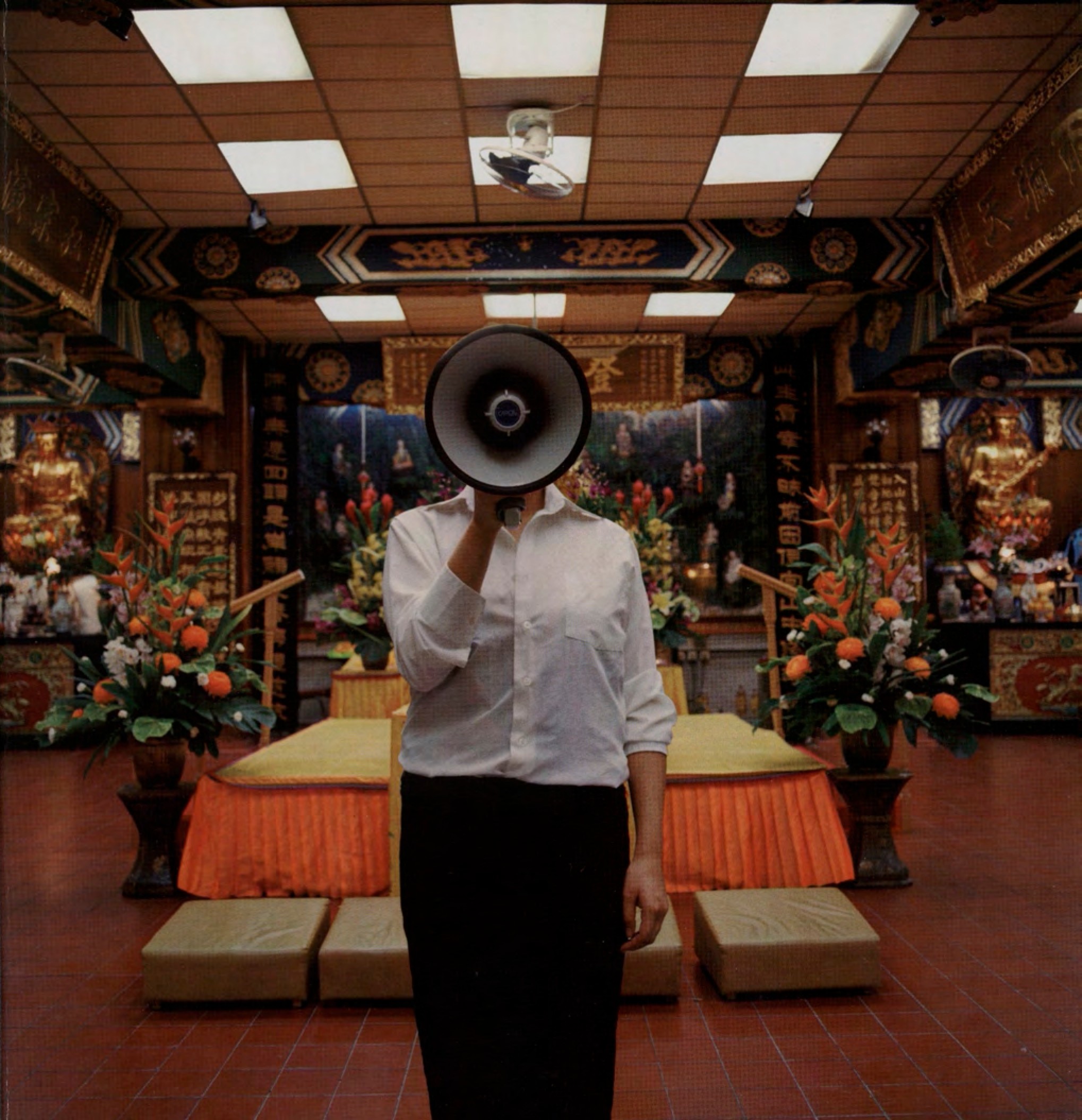 A woman stands among flowers and statues, speaking into a megaphone.