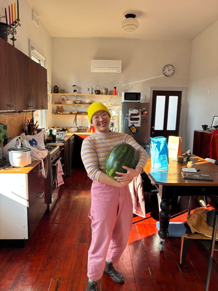 A woman in a yellow beanie and pink pants stands in a kitchen holding a large watermelon