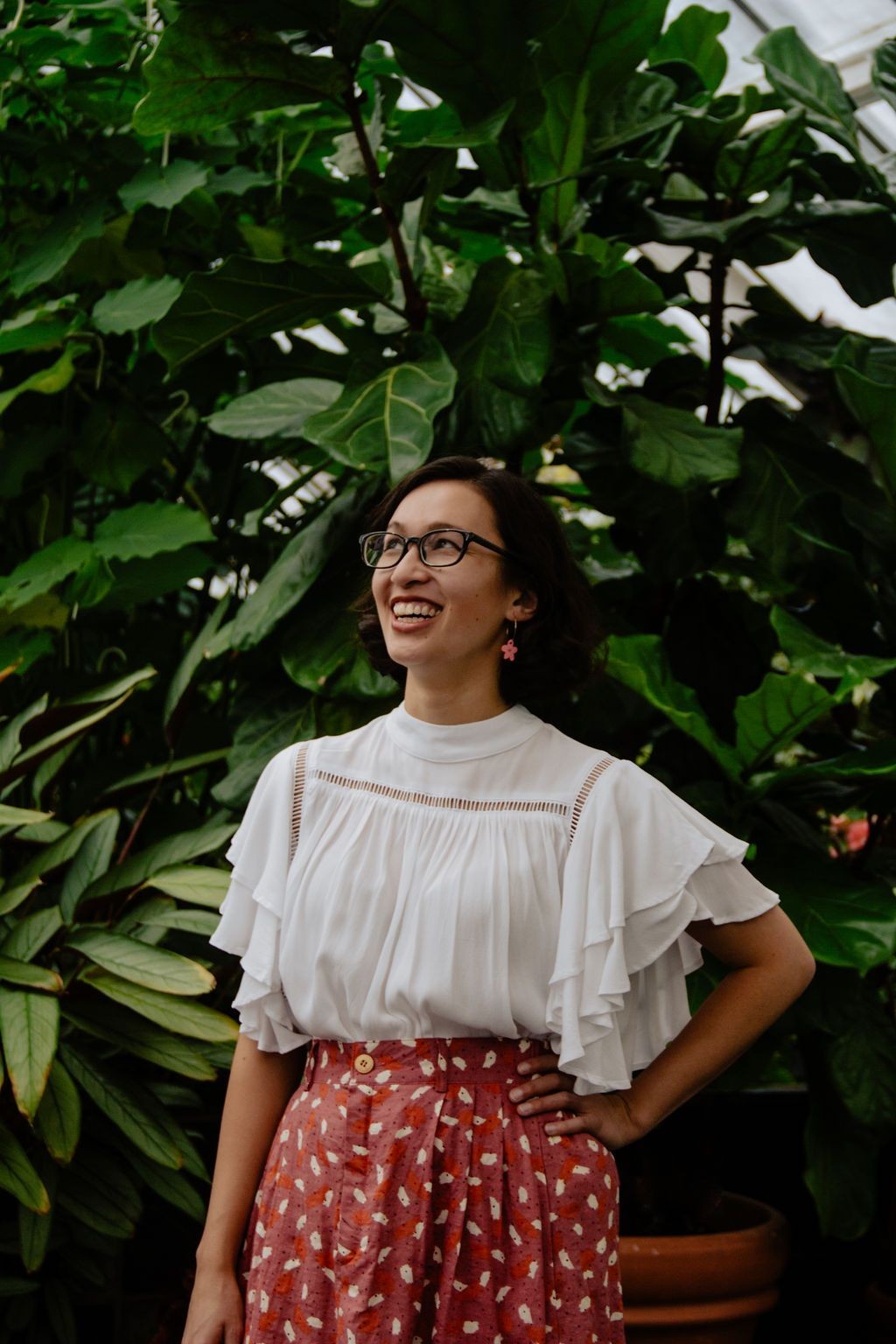 A woman with glasses and a black bob, wearing a white frilly top and orange polka dot skirt, stands in front of foliage