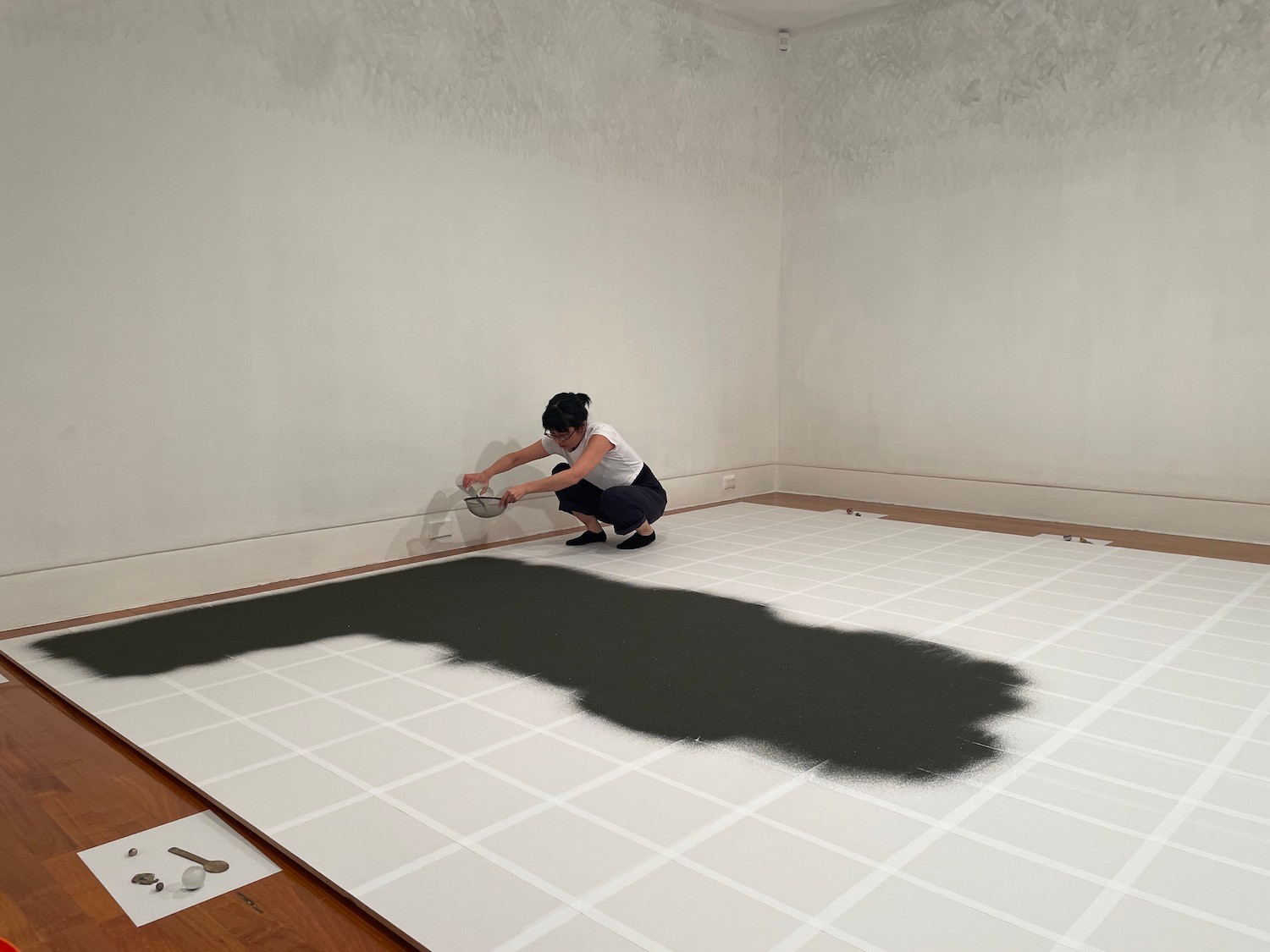 The artist crouching near the wall and scattering black sand onto the floor that has been gridded with white paper.