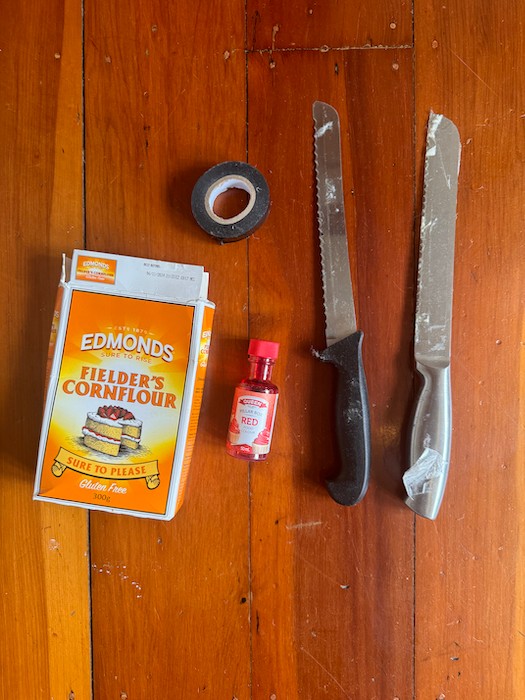 A box of cornflour, a bottle of food colouring, a roll of electrical tape and two knives are laid on the floor.