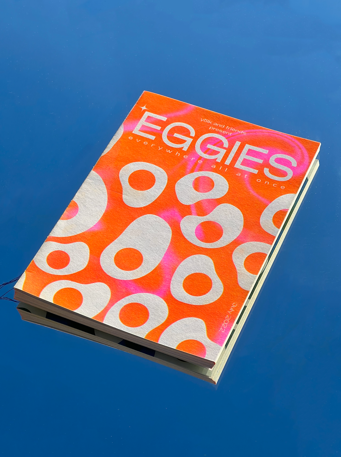 A bright orange zine with abstract egg patterns, photographed on a blue background. 