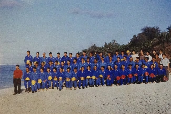 Three rows of Chinese workers in blue overalls pose for a group photo on a beach in Rarotonga.