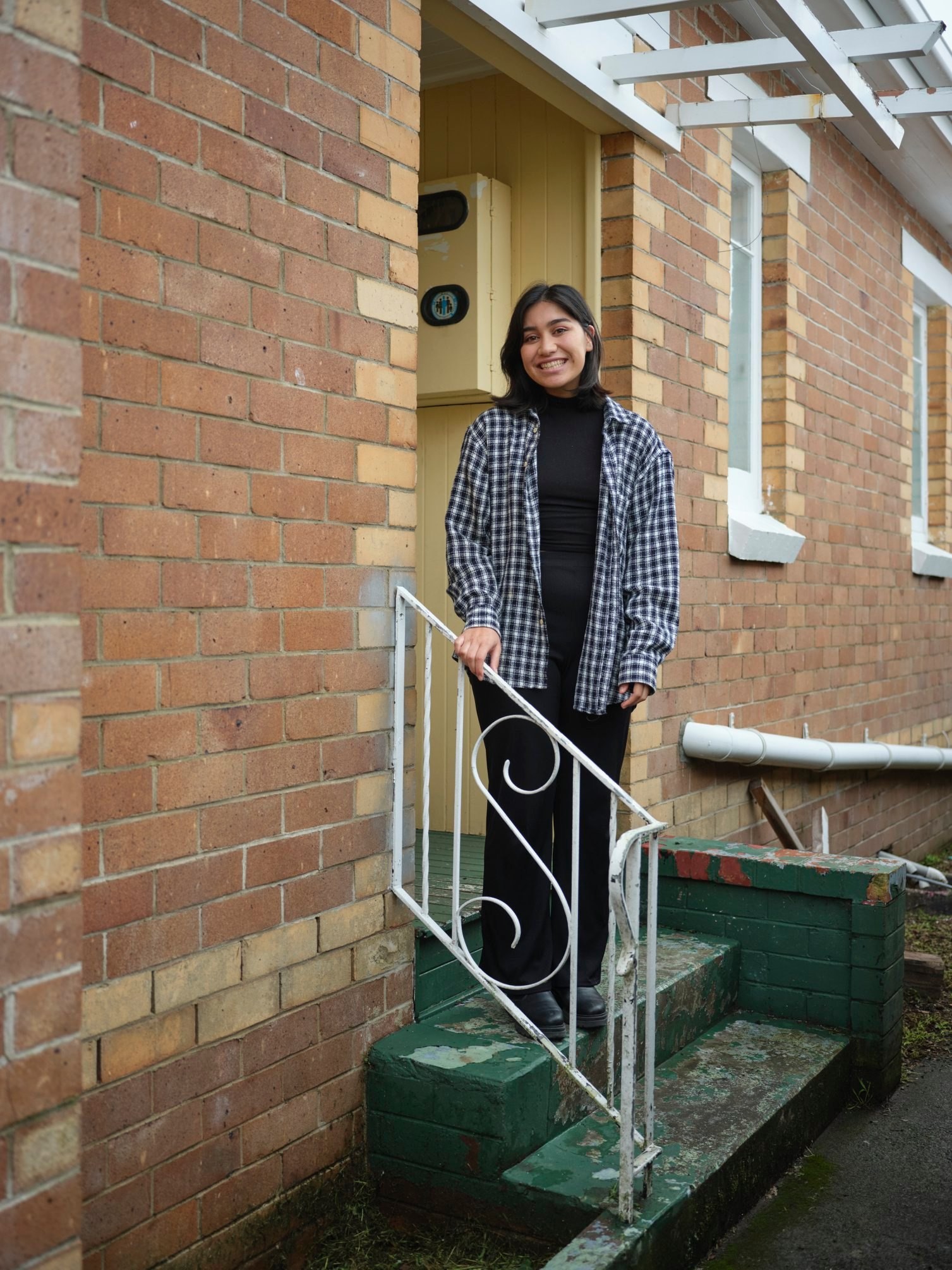 Person with shoulder length hair wearing a blue checkered top and smiling while posing in front of a house entryway