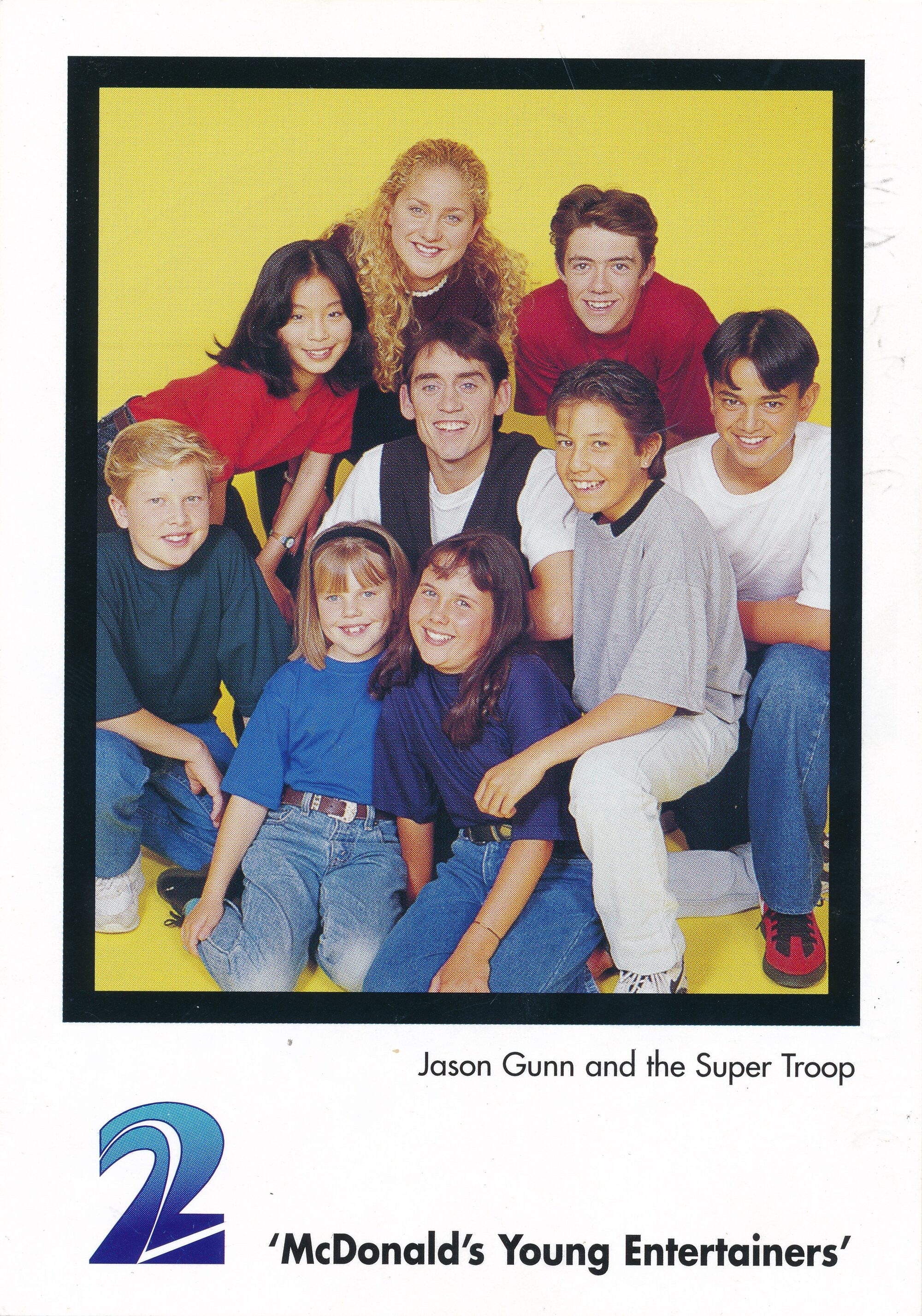 A group of smiling children and teens, including host Jason Gunn, pose together in front of a bright yellow background for the TV show 'McDonald’s Young Entertainers'.