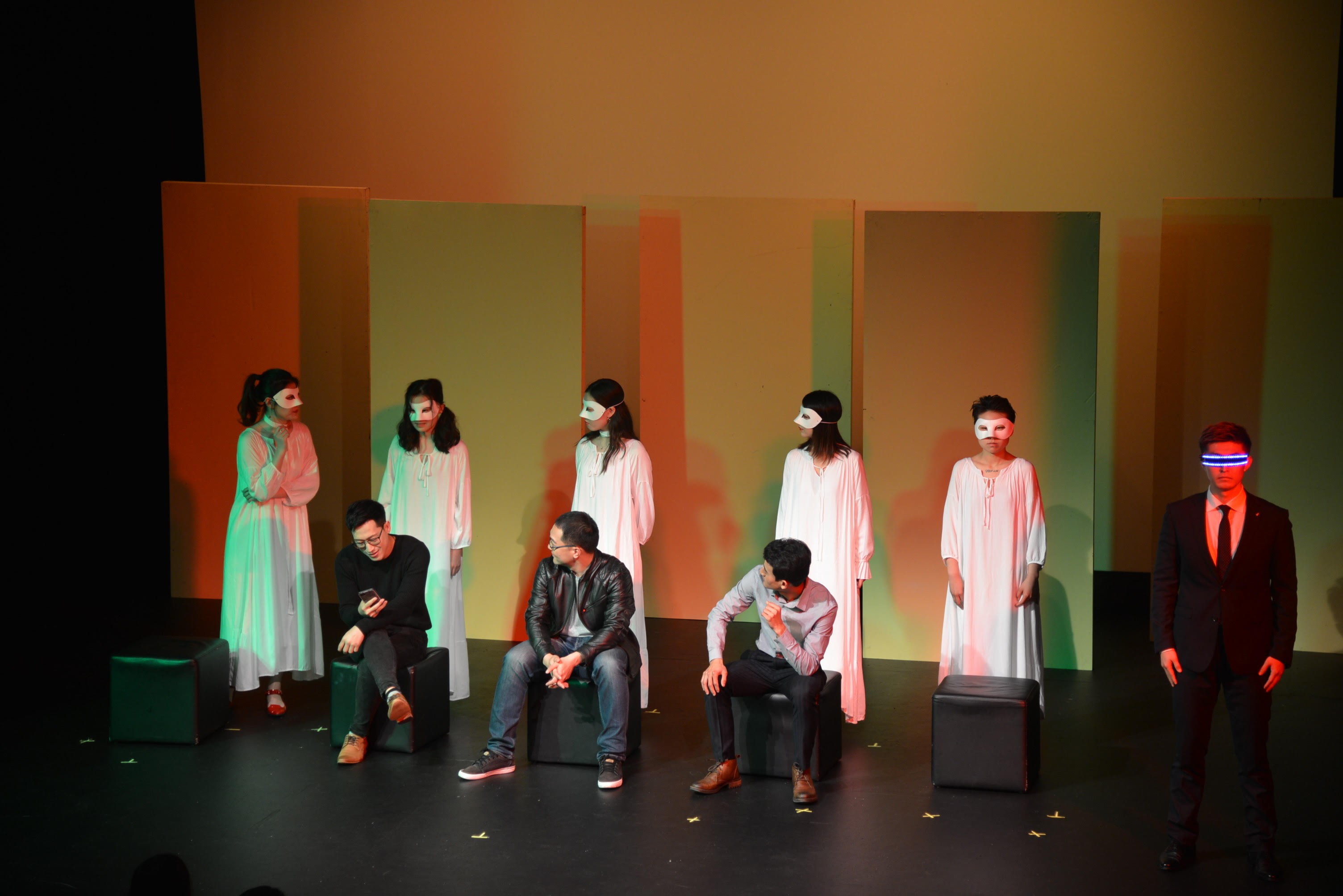 Five women in long white dresses and white masks stand in the background while three men in casual clothing sit on blocks in front of them. At the front of a stage, a man wearing a suit and holographic glasses stares into the audience