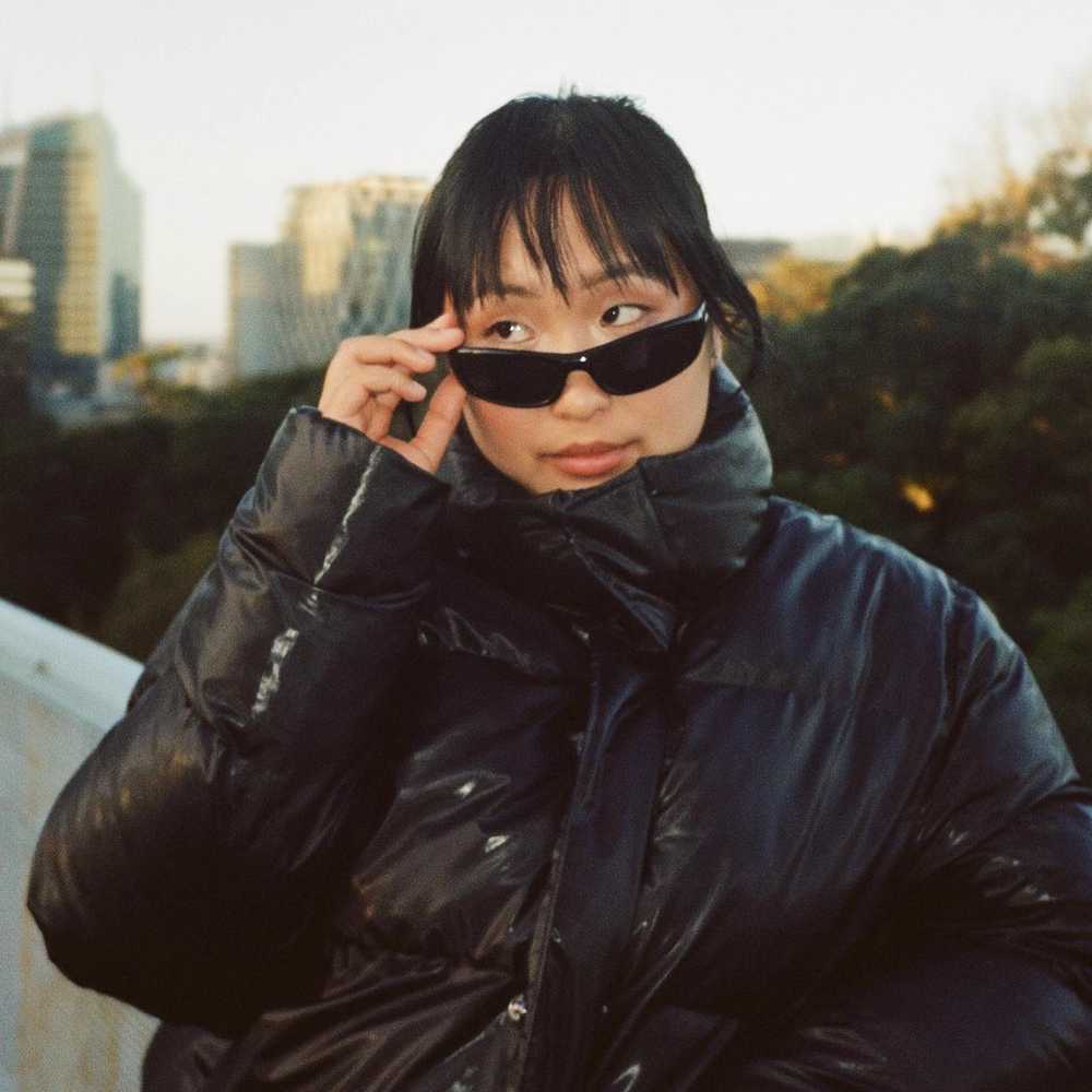 Jess Fu has a fringe and wears a black puffer jacket. She pulls down her sunglasses to look over them.