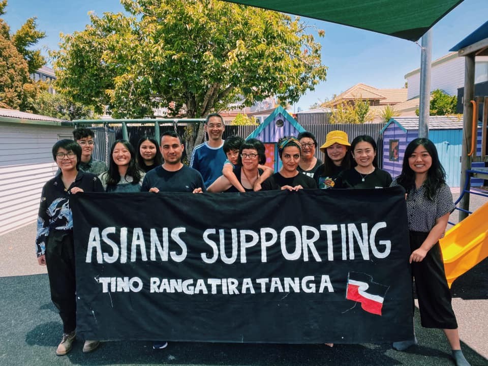 13 people grouped around a banner saying 'Asians Supporting Tino Rangatiratanga' with the Tino Rangatiratanga flag painted on