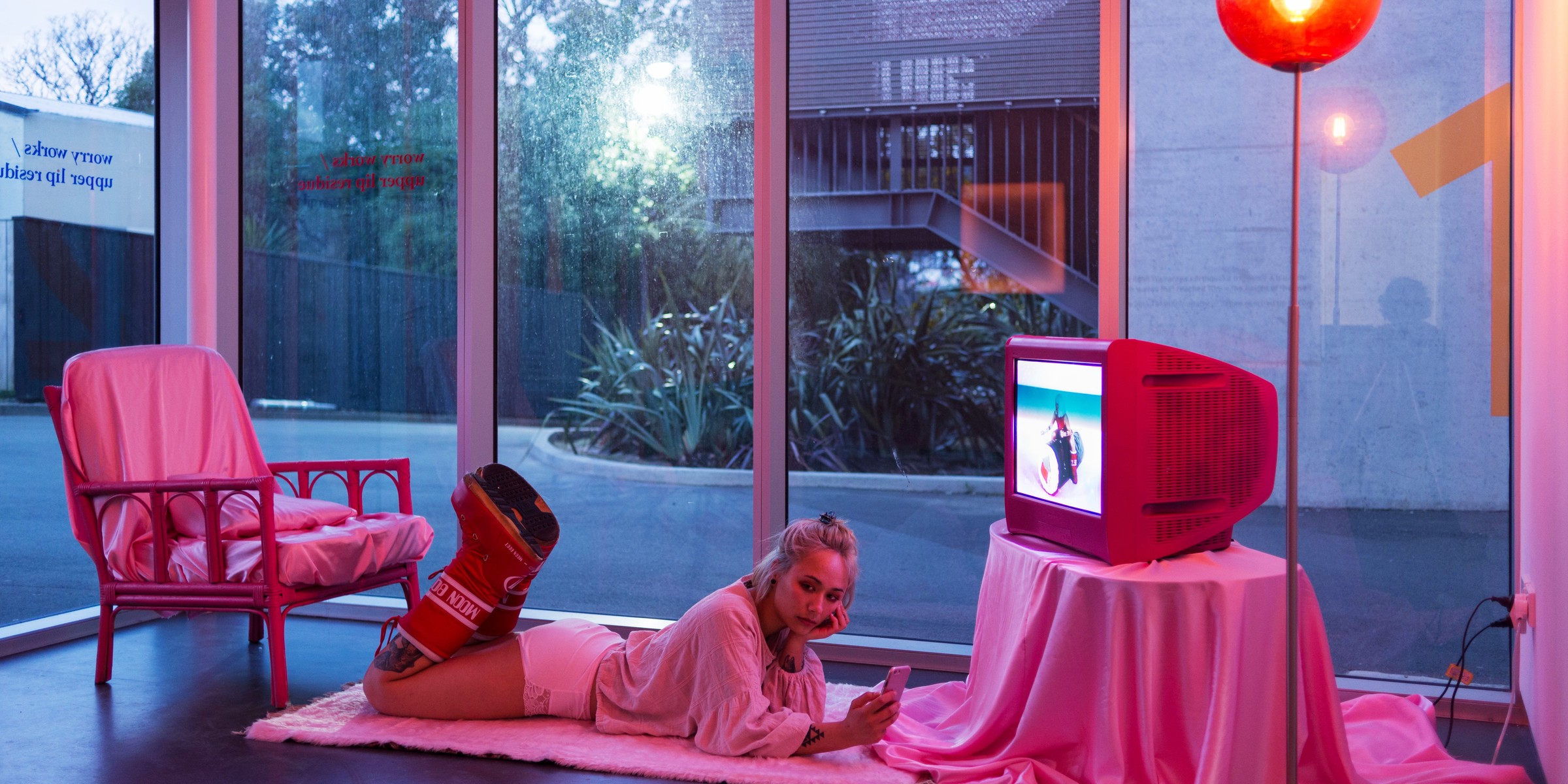 In a glass gallery filled with pink light, Emiko Sheehan lies on a small rug, with a TV in front of her.