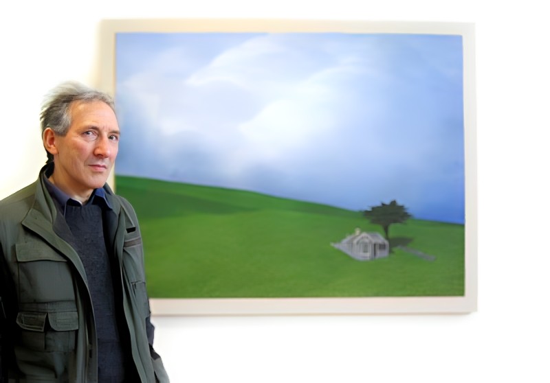 Brent Wong stands in a jacket with lots of pockets, in front of a painting showing an expanse of cloud-ed sky, a rolling hill and a small house near a tree.