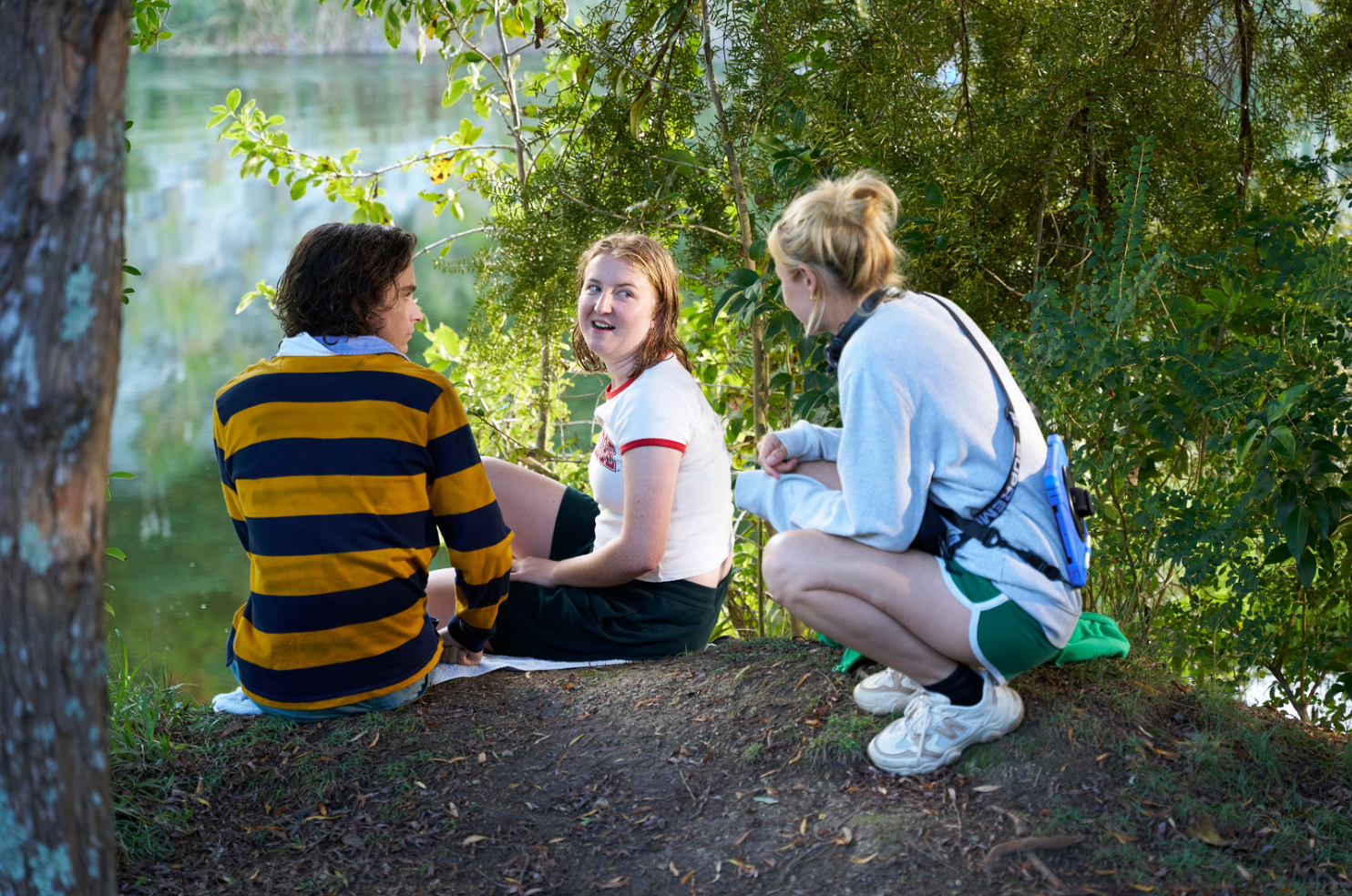 A woman with bleached hair and a grey sweatshirt talks to two actors sitting by the trees