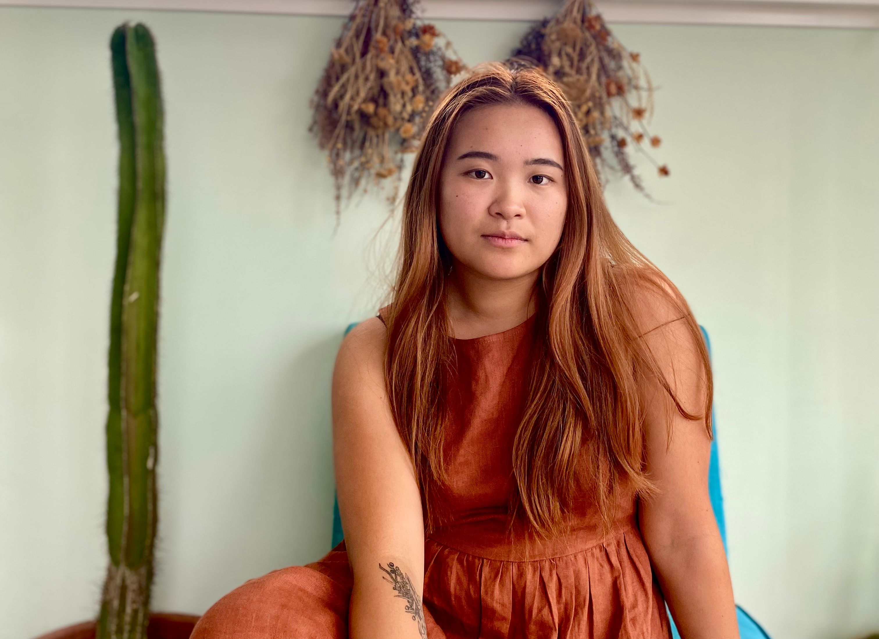 Eda has long hair with copper highlights, and poses in front of some dried flowers for the photo.