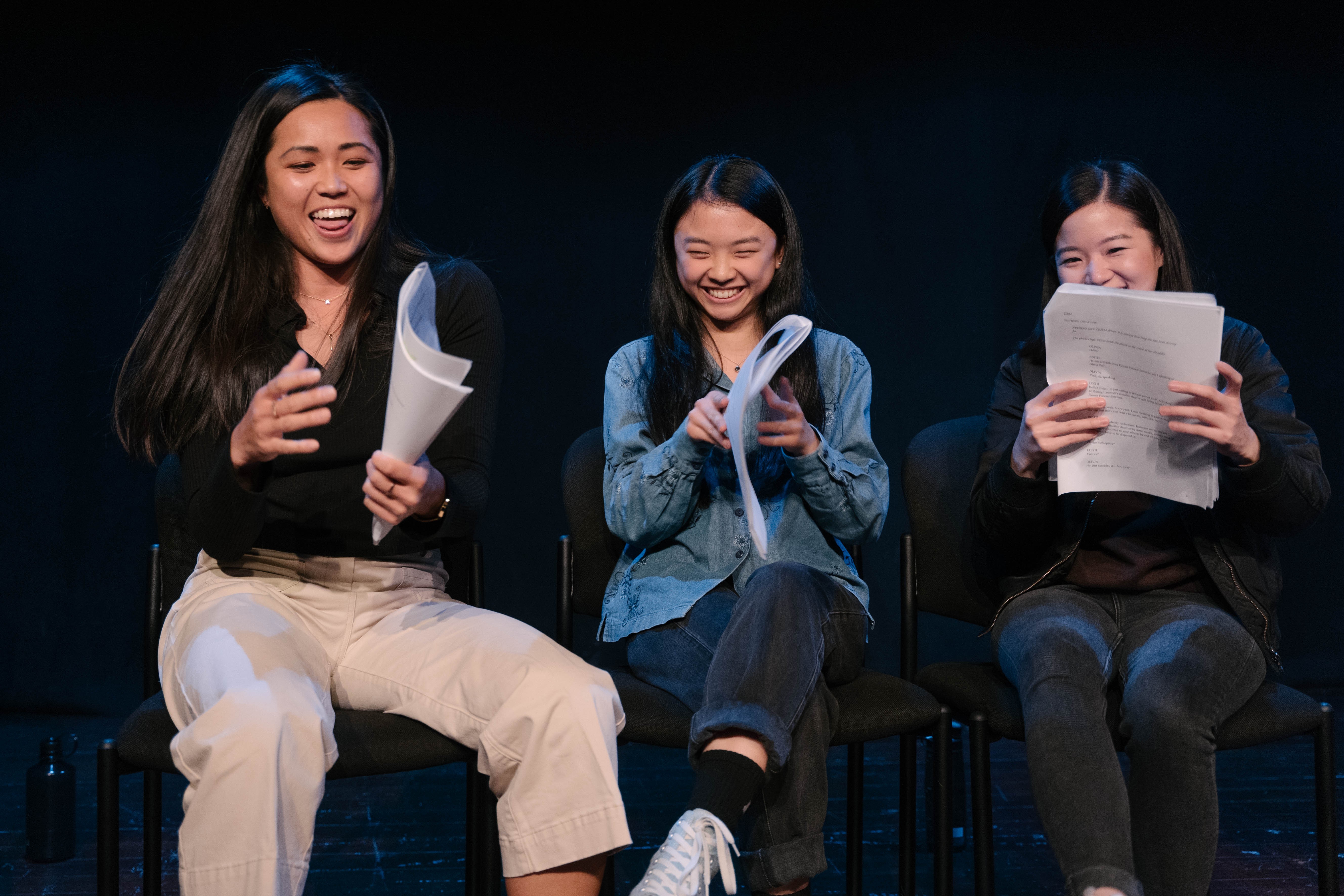 Three East asian woman sit on chairs holding their scripts and laughing