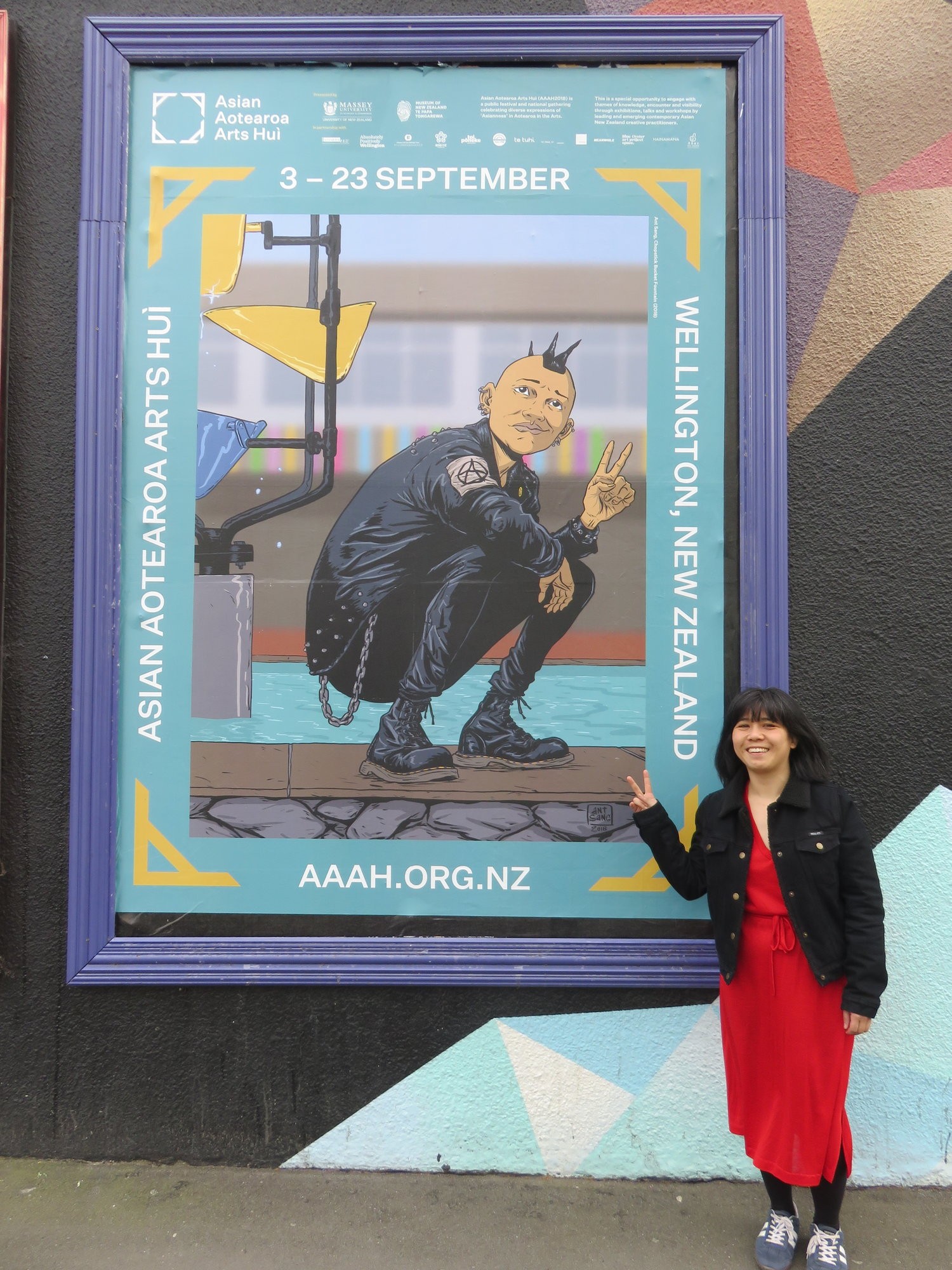 A smiling Kerry Ann Lee does the peace sign with her fingers in front of a large poster for the hui, featuring the work of Ant Sang.