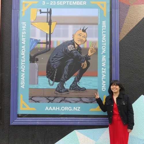 A smiling Kerry Ann Lee does the peace sign with her fingers in front of a large poster for the hui, featuring the work of Ant Sang.