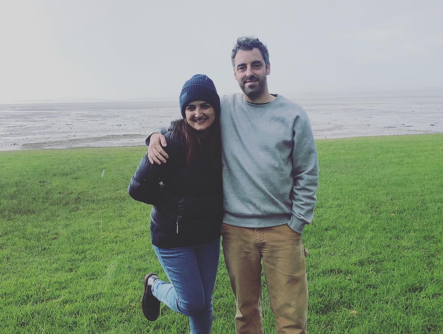 A woman and man stand arm-in-arm on a grassy field by the shore, smiling at the camera on an overcast day.