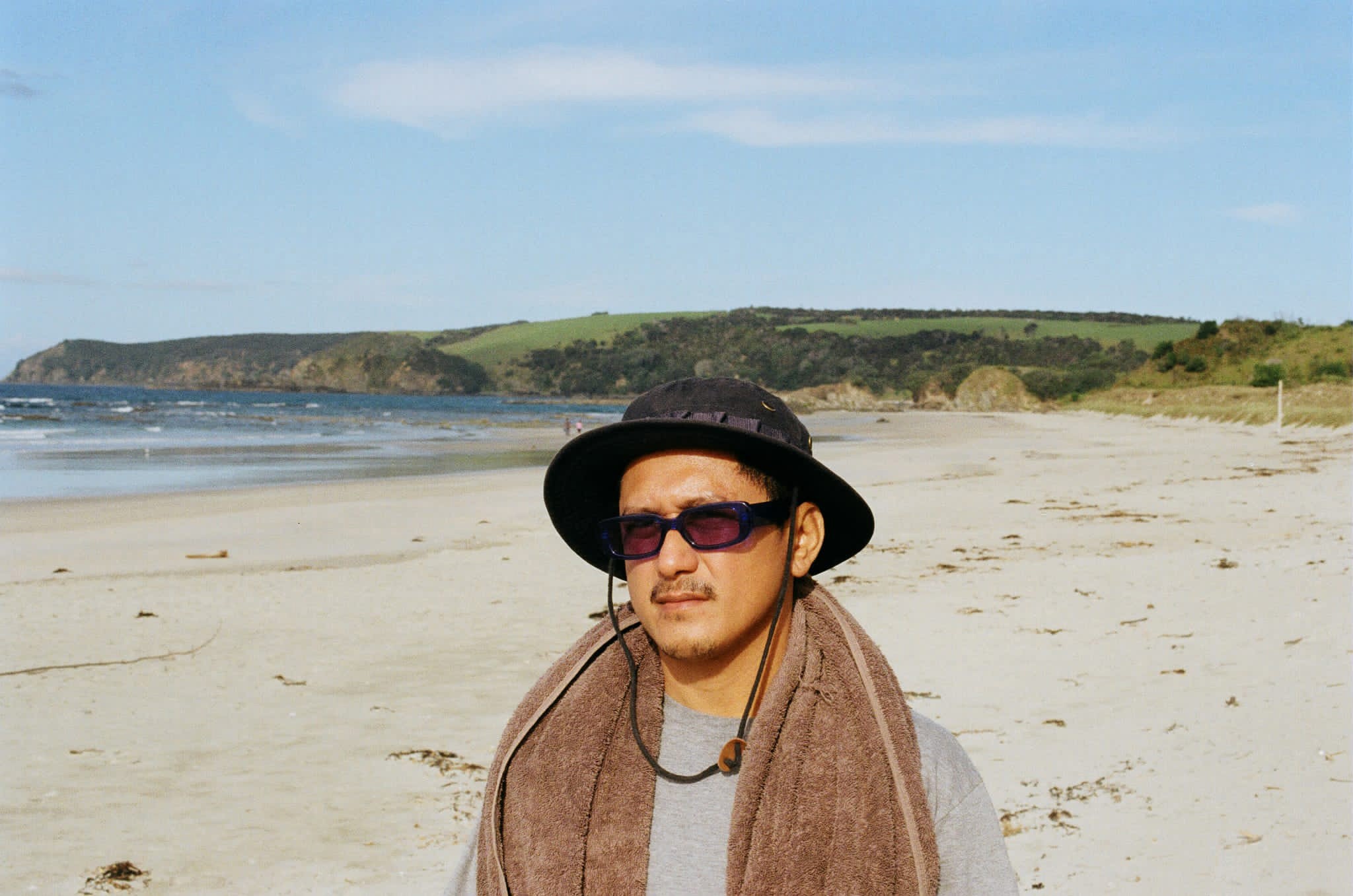 Person of Filipino descent wearing dark sunnies, wide brimmed hat with a towel around their shoulders at the beach