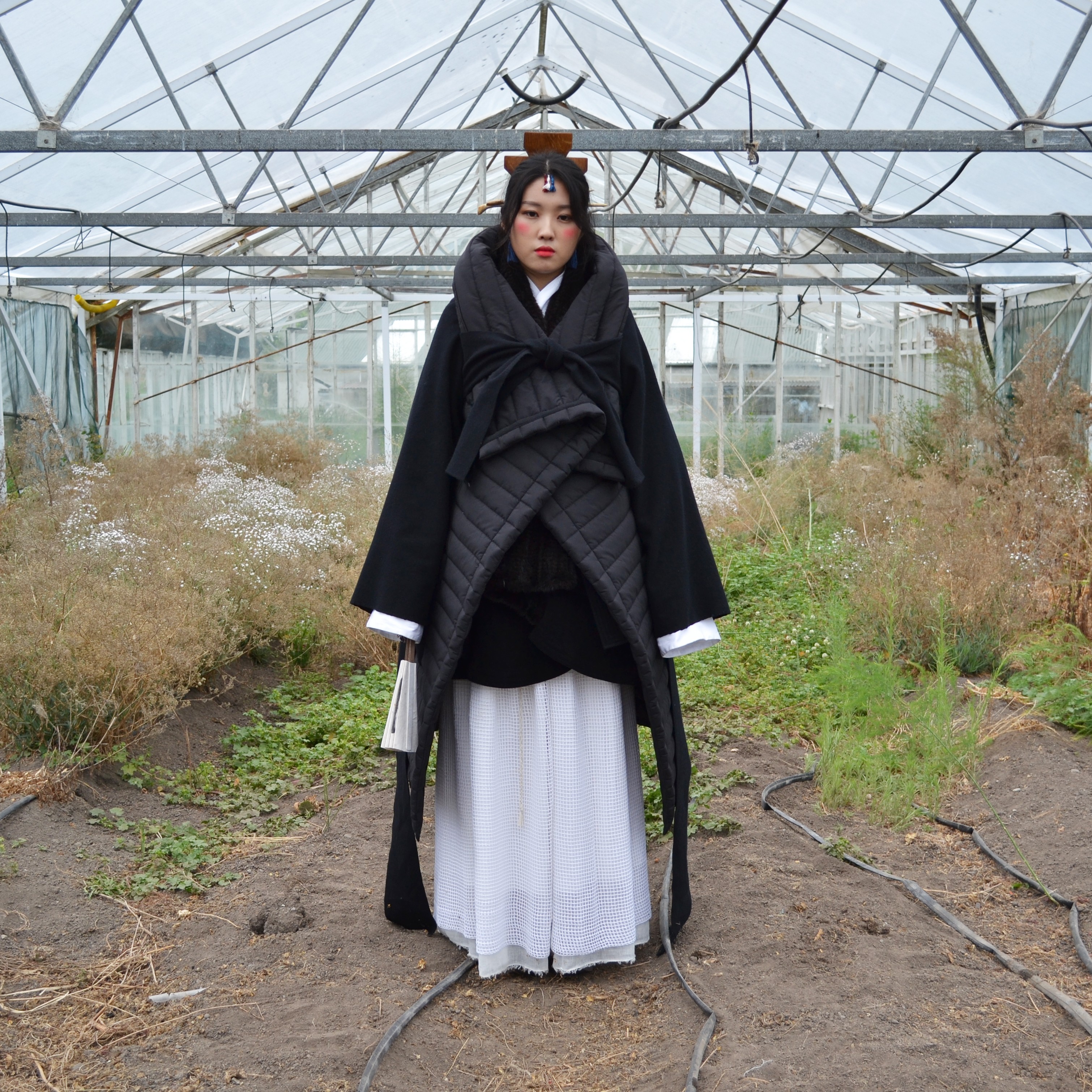 A woman in flowing hanbok-inspired garments stands in an abandoned greenhouse.