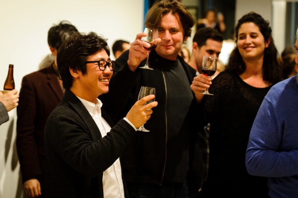 Seung Yul Oh smiling and raising a glass amongst a group of poeple, with a man and a woman next to him similarly raising their glasses. 