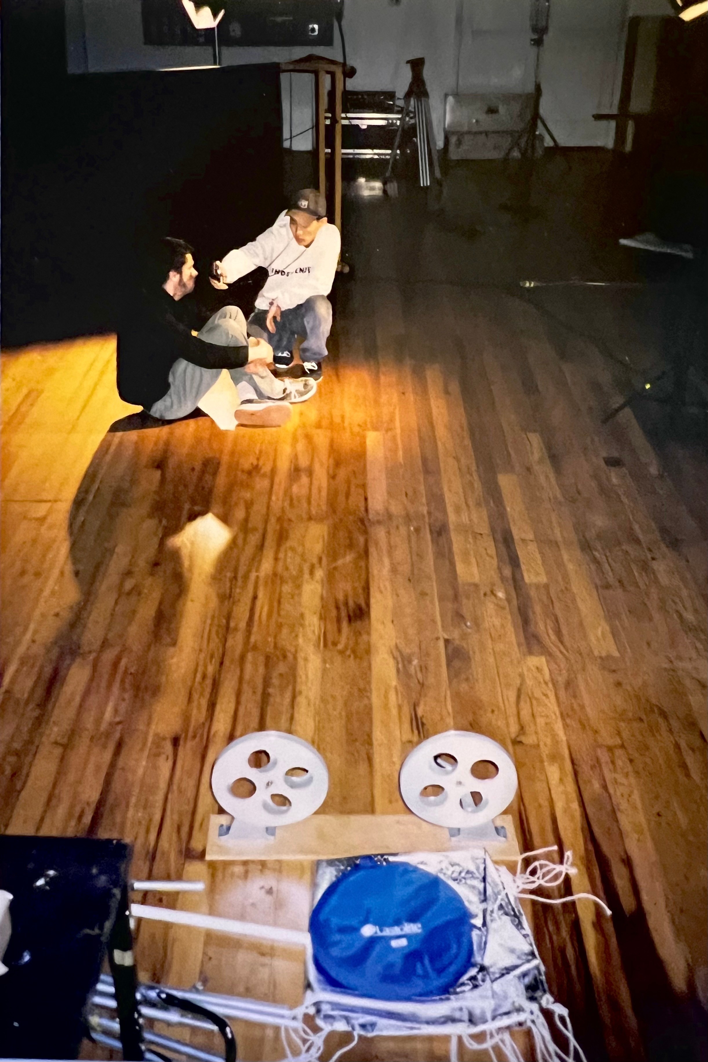 A man in a black top and grey shorts sits on the floor next to a man in a white hooded sweatshirt and black baseball cap. In the foreground are various items of film equipment.